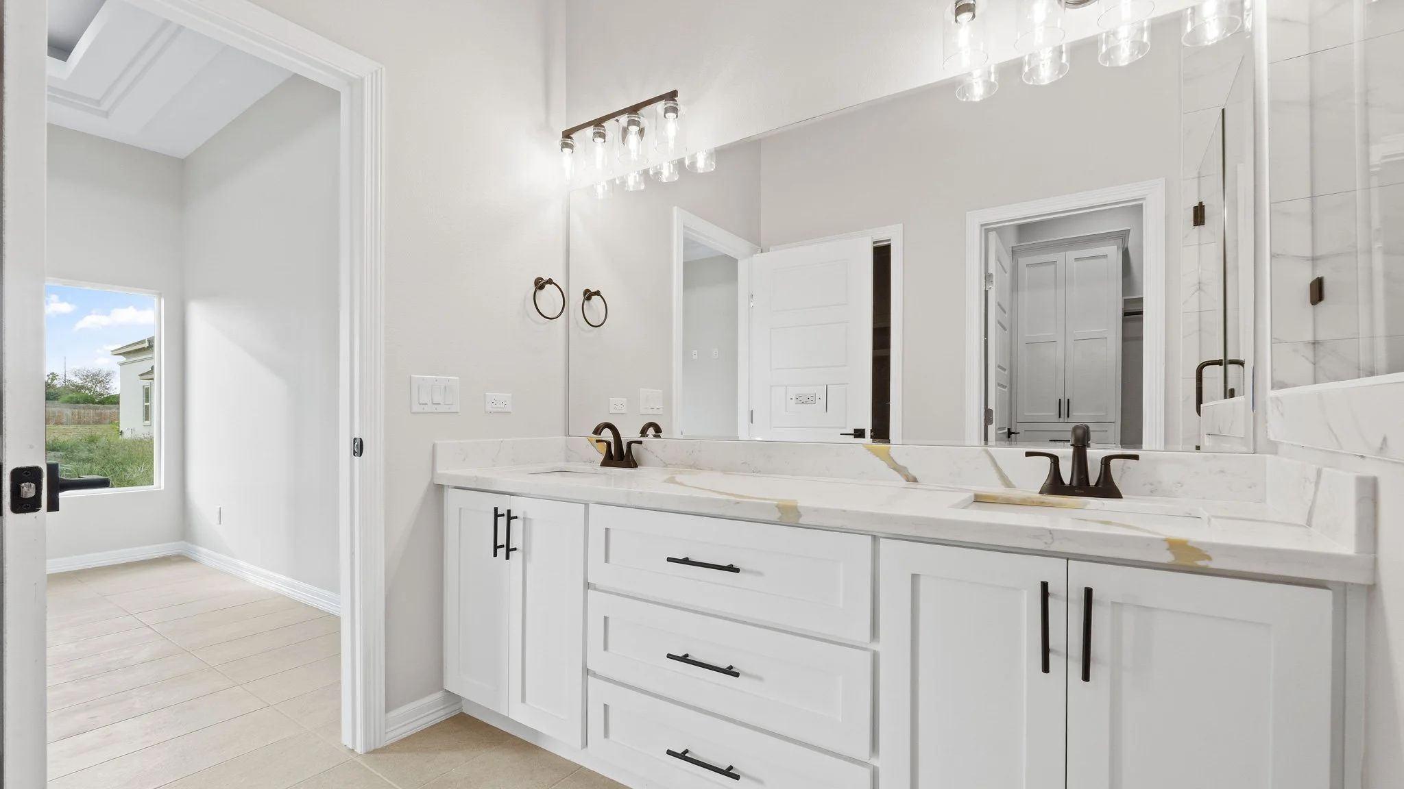 Lavabo de baño con espejo grande, encimera de mármol blanco, lavabo doble con grifos en acabado negro, y armarios blancos, en baño moderno con paredes blancas y un accesible ventanal hacia el exterior.