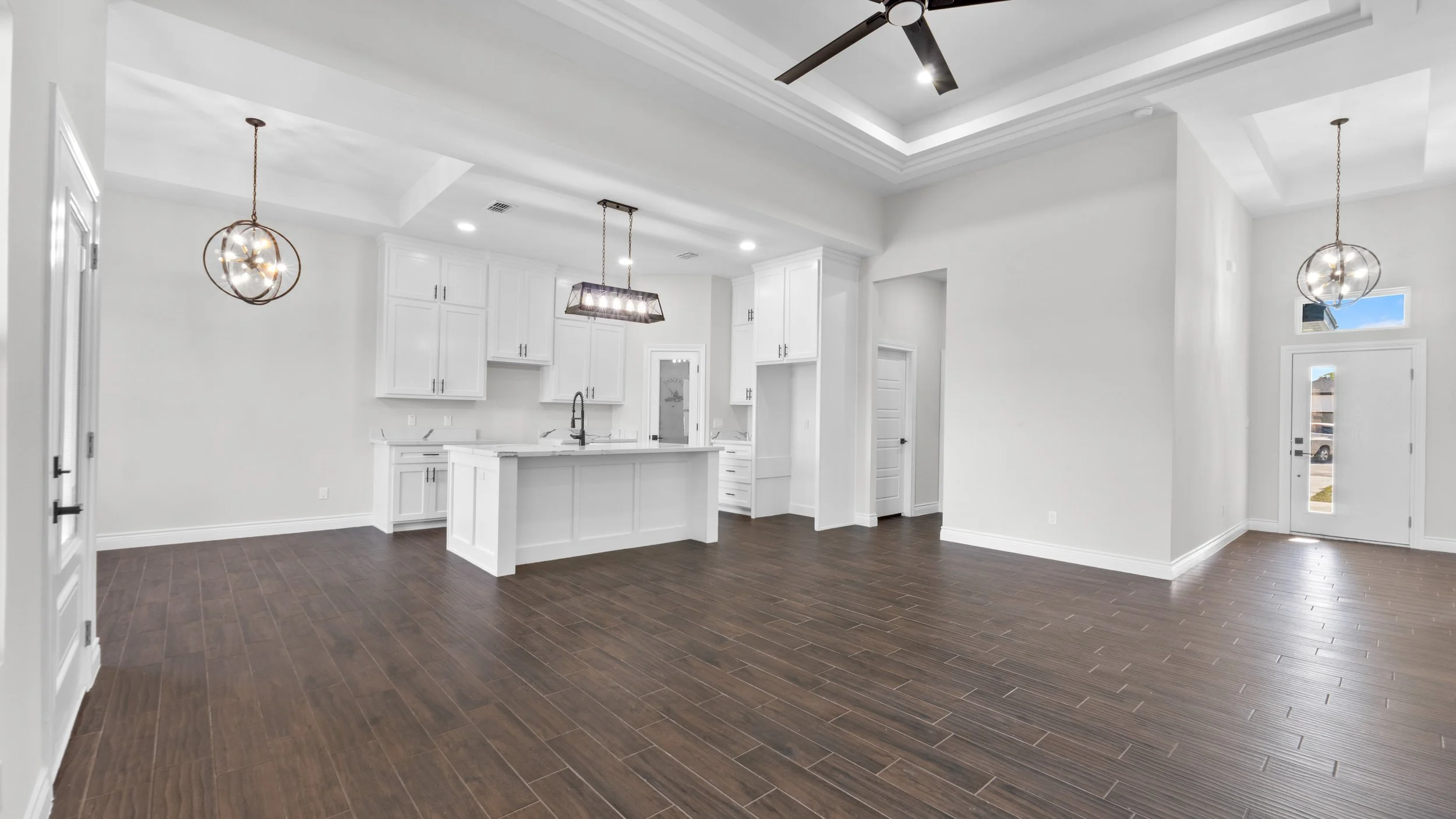 Interior de una sala y cocina moderna y luminosa con paredes blancas, suelo de madera oscura y techos altos con iluminación empotrada y ventilador.