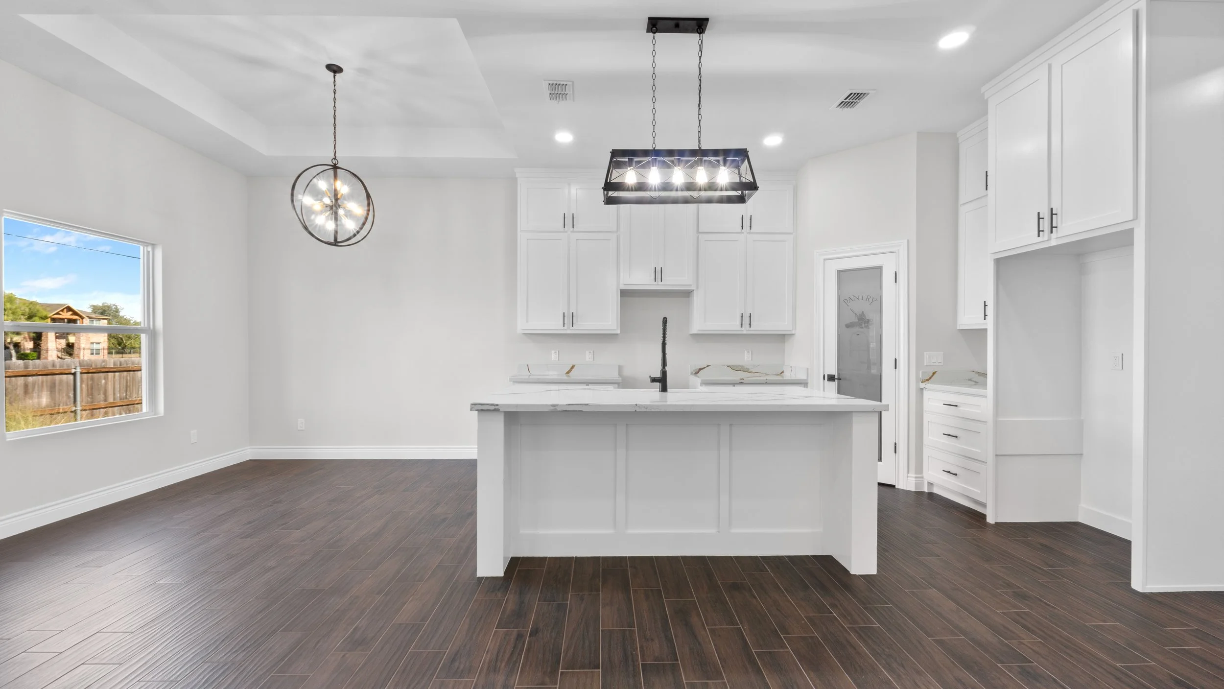 Cocina moderna con encimera de mármol, gabinetes blancos, lámpara rectangular y lámpara de techo redonda, suelo de madera oscura, ventana grande con vistas a un patio.