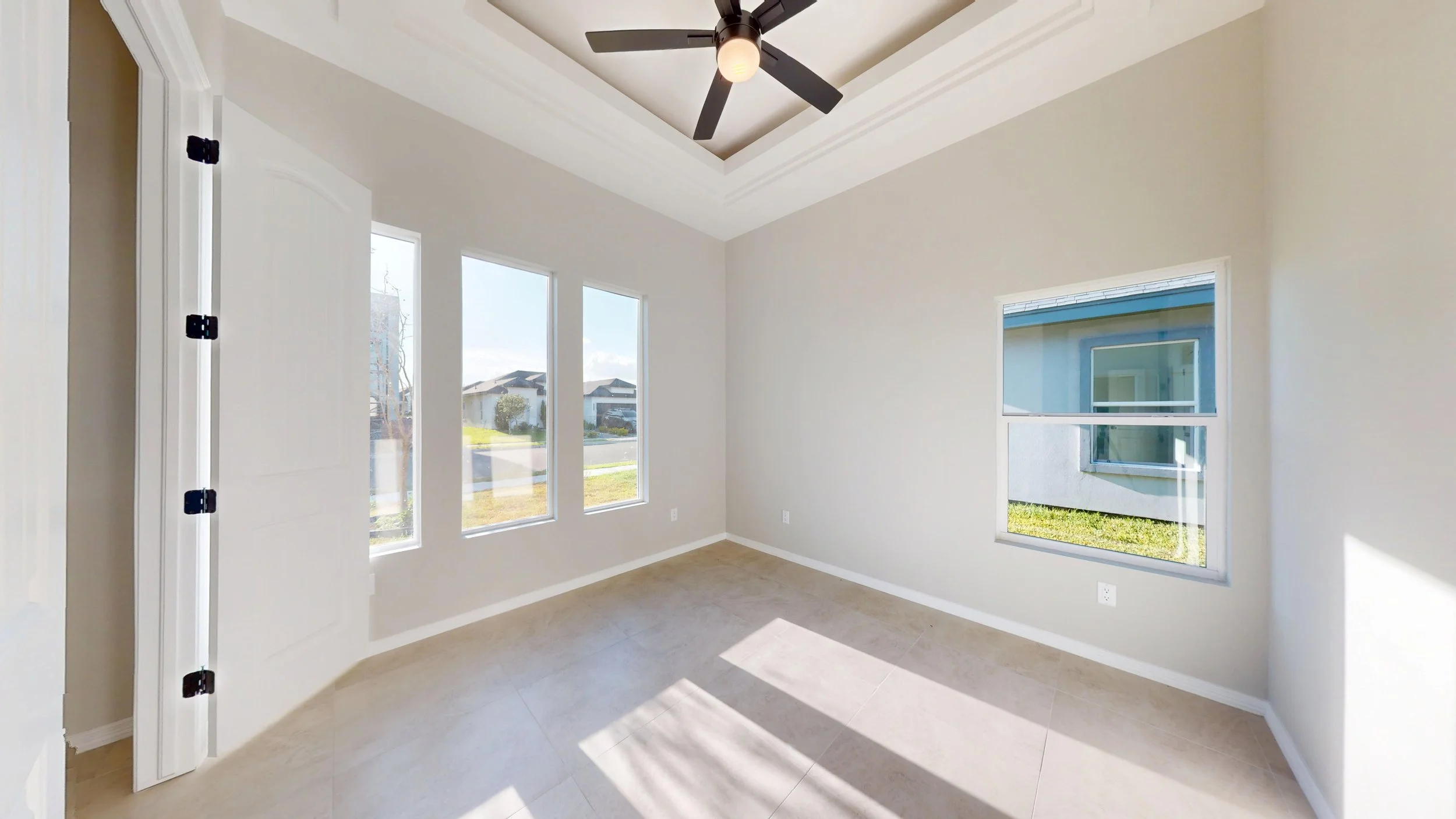 Habitación vacía con paredes blancas, ventanas grandes con vista al exterior, techo con ventilador moderno y suelo de baldosas claras.