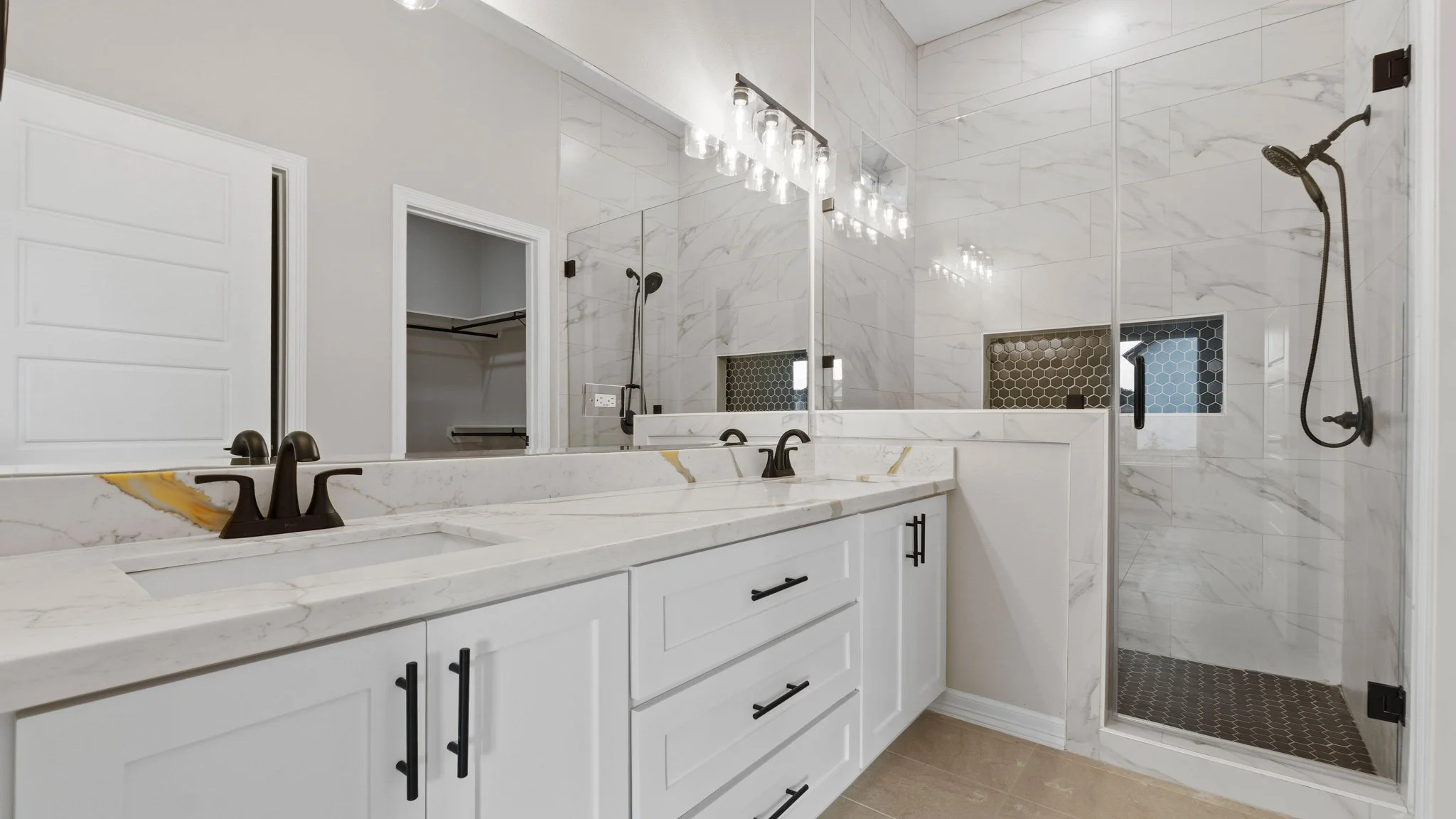 Lavabo de doble seno con encimera de mármol blanco, grifos negros, large espejo y luz de focos, ducha de cristal con azulejos de mármol y nichos con azulejos de mosaico en la pared del baño