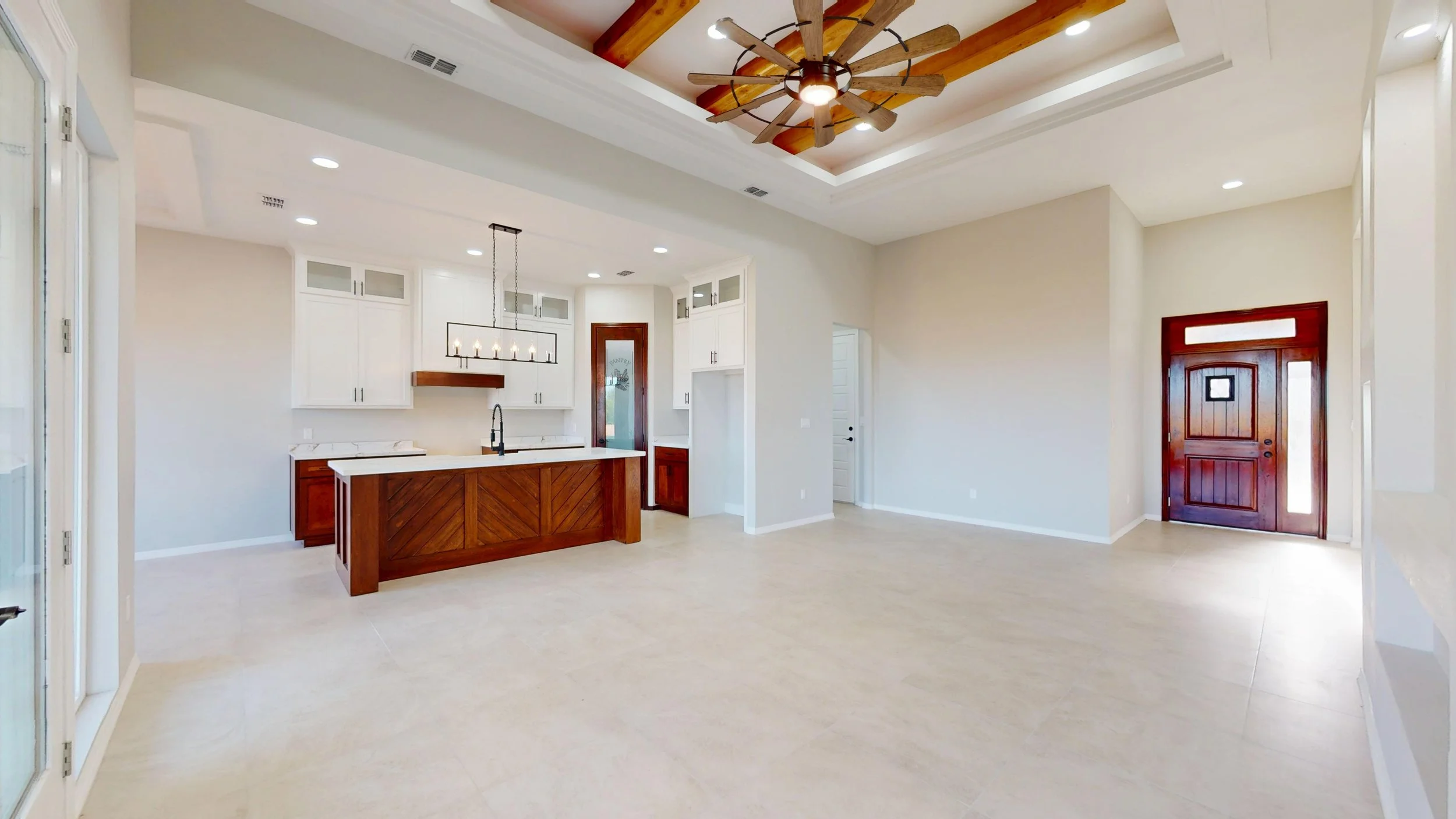 Espaciosa sala de estar en una casa moderna con suelo de baldosas beige, paredes blancas, puerta de madera al fondo y cocina con isla de madera y gabinetes blancos a la izquierda. Techos altos con detalles en madera y ventilador de techo grande.