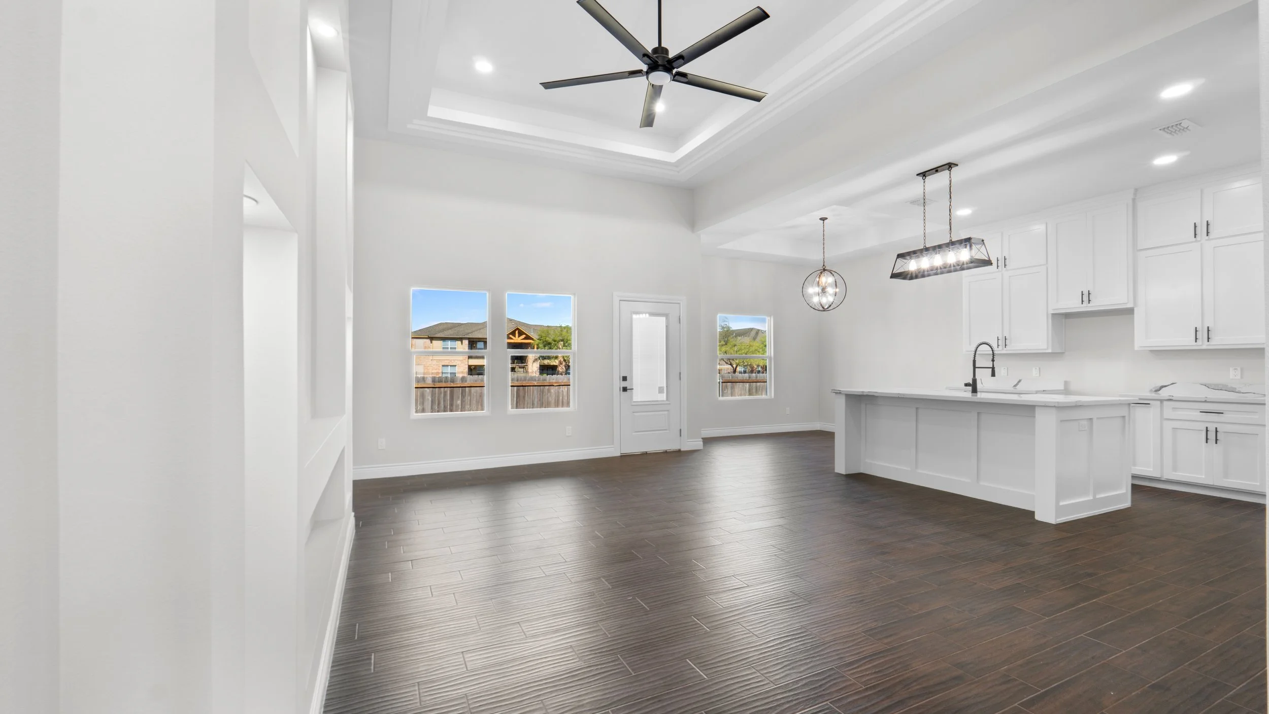 Espacio abierto de cocina y comedor con ventanas, puertas de cristal, suelo de madera oscura, paredes y muebles blancos, lámparas modernas y ventilador en el techo