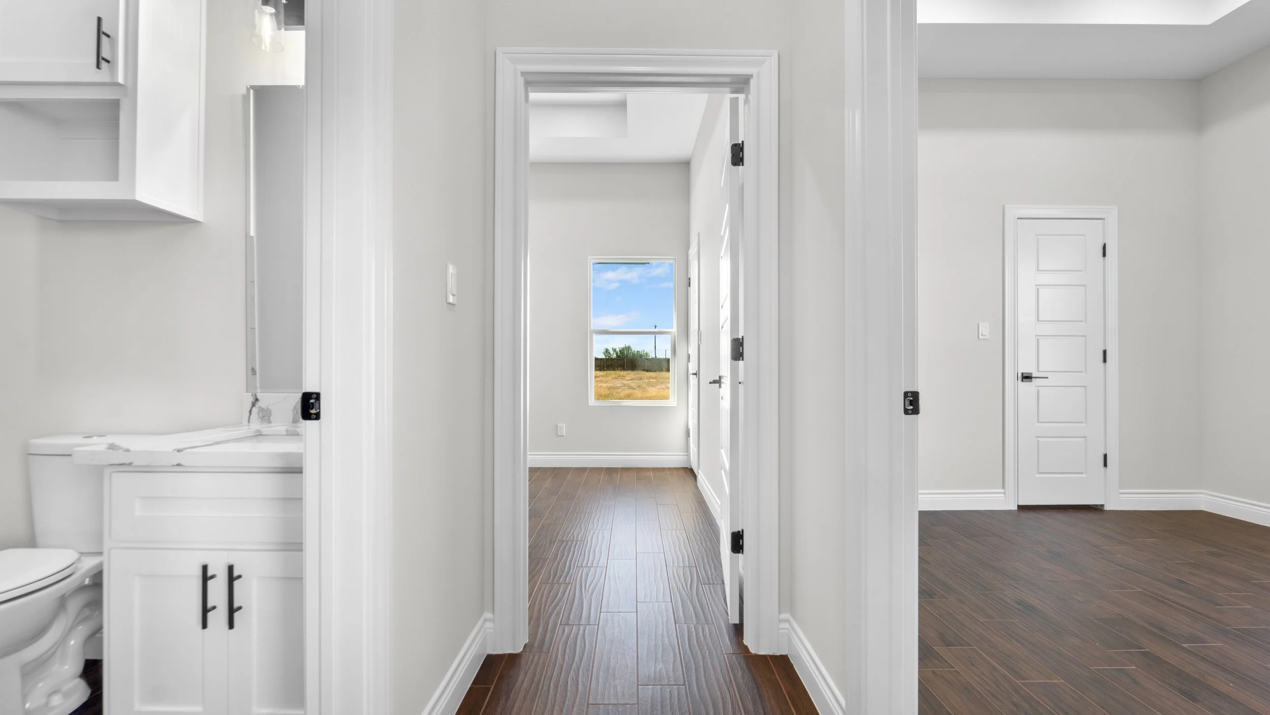 Vista de diferentes habitaciones con suelos de madera en una casa moderna, con paredes blancas y puertas blancas, y una ventana que muestra un paisaje exterior con cielo azul y nubes.