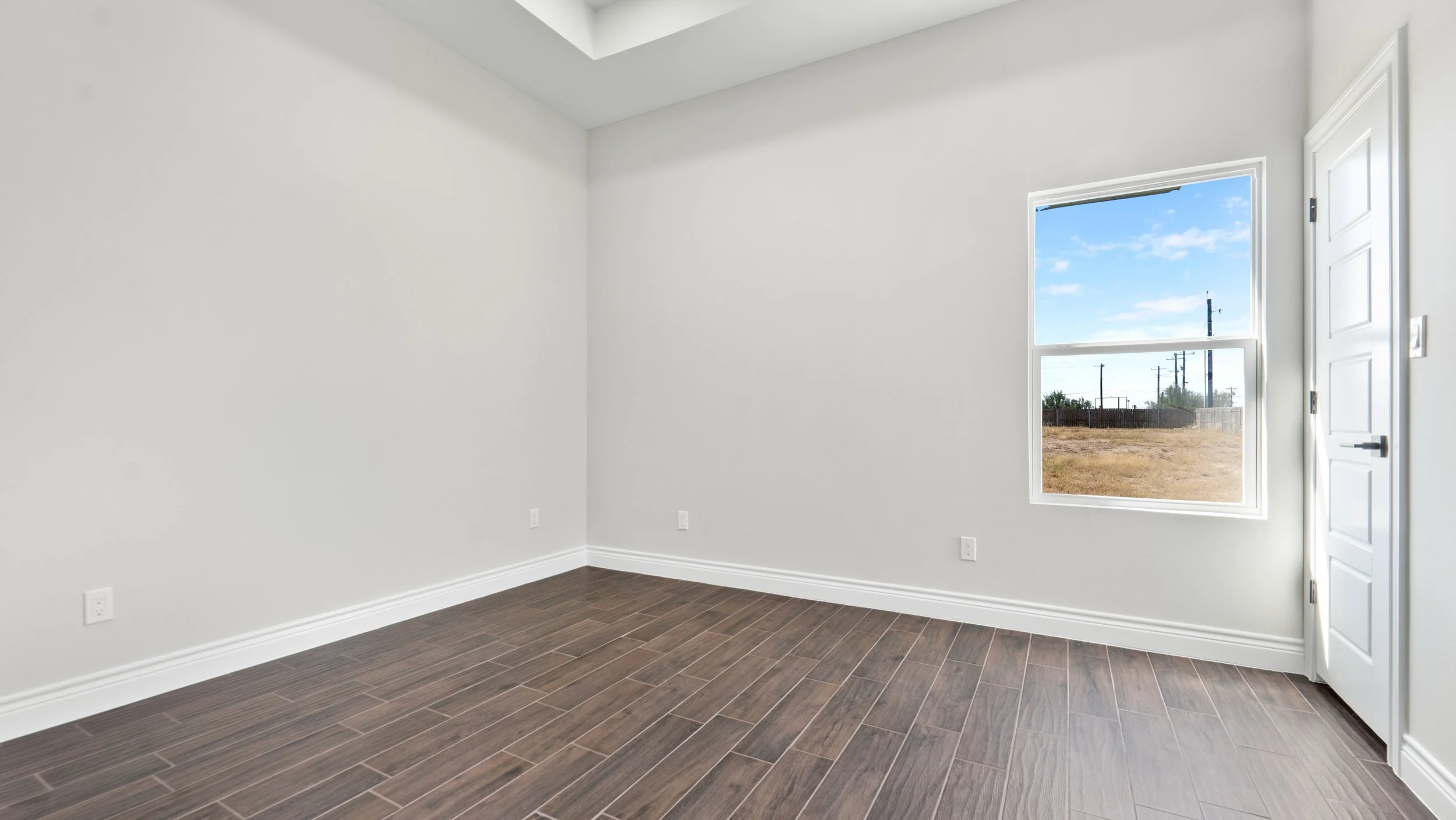 Cuarto vacío con paredes blancas, piso de madera oscura y una ventana con vista al exterior