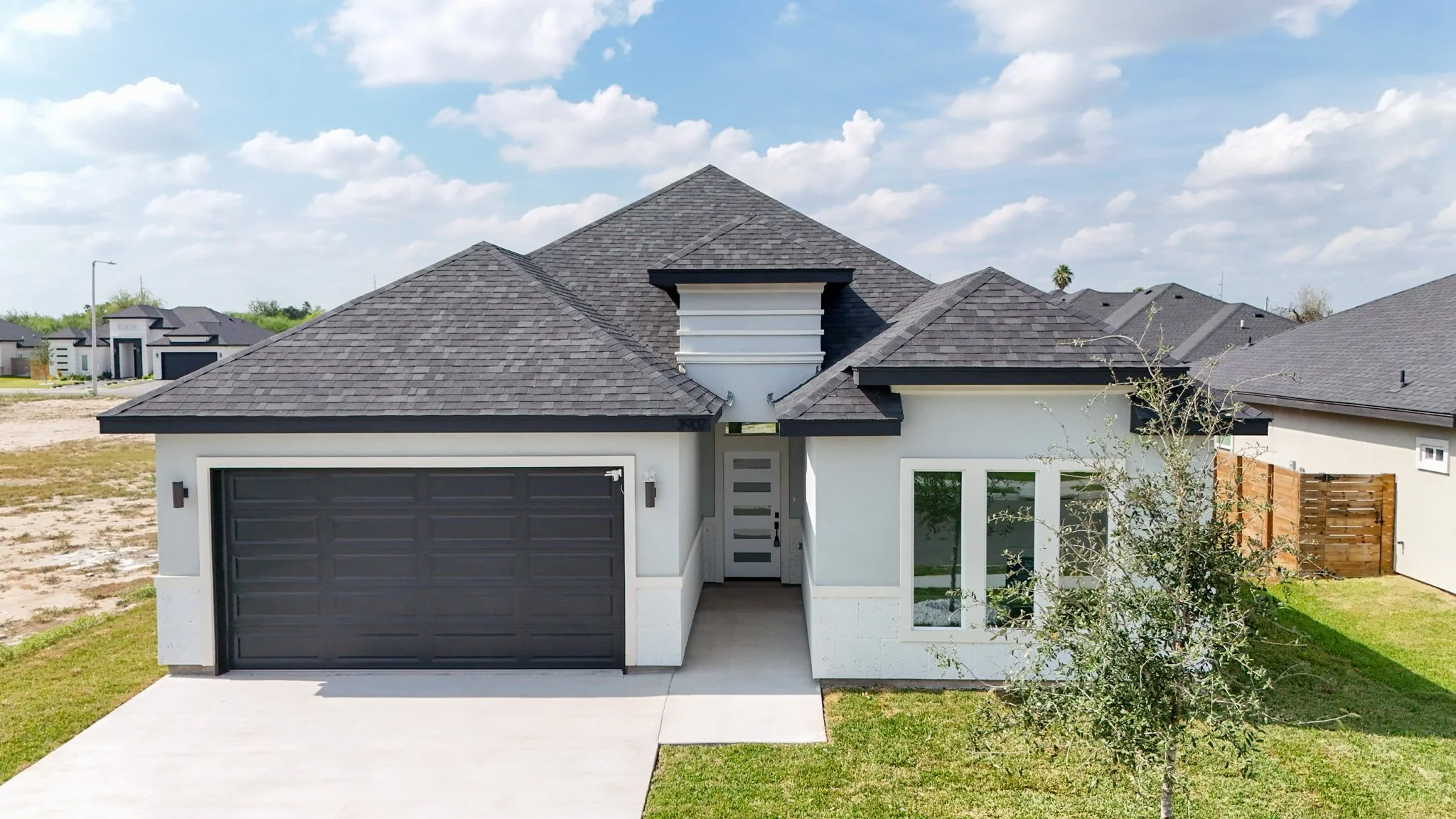 Casa moderna con techo de tejas grises, garaje doble y ventanas grandes, en vecindario con cielo azul y nubes esponjosas.