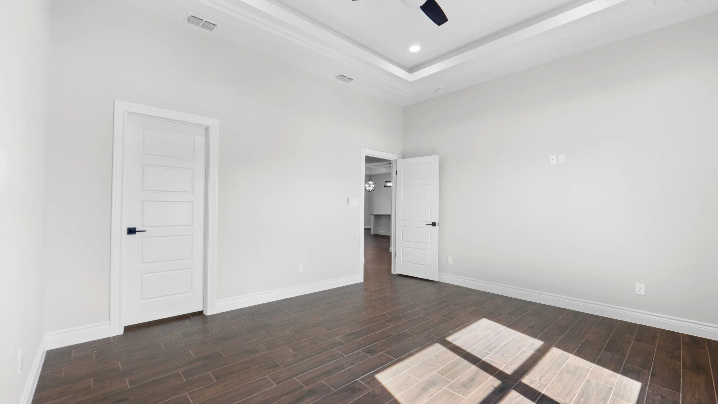 Habitación vacía con paredes blancas, suelo de tablones de madera oscura, luz natural entrando por una ventana y puertas blancas.
