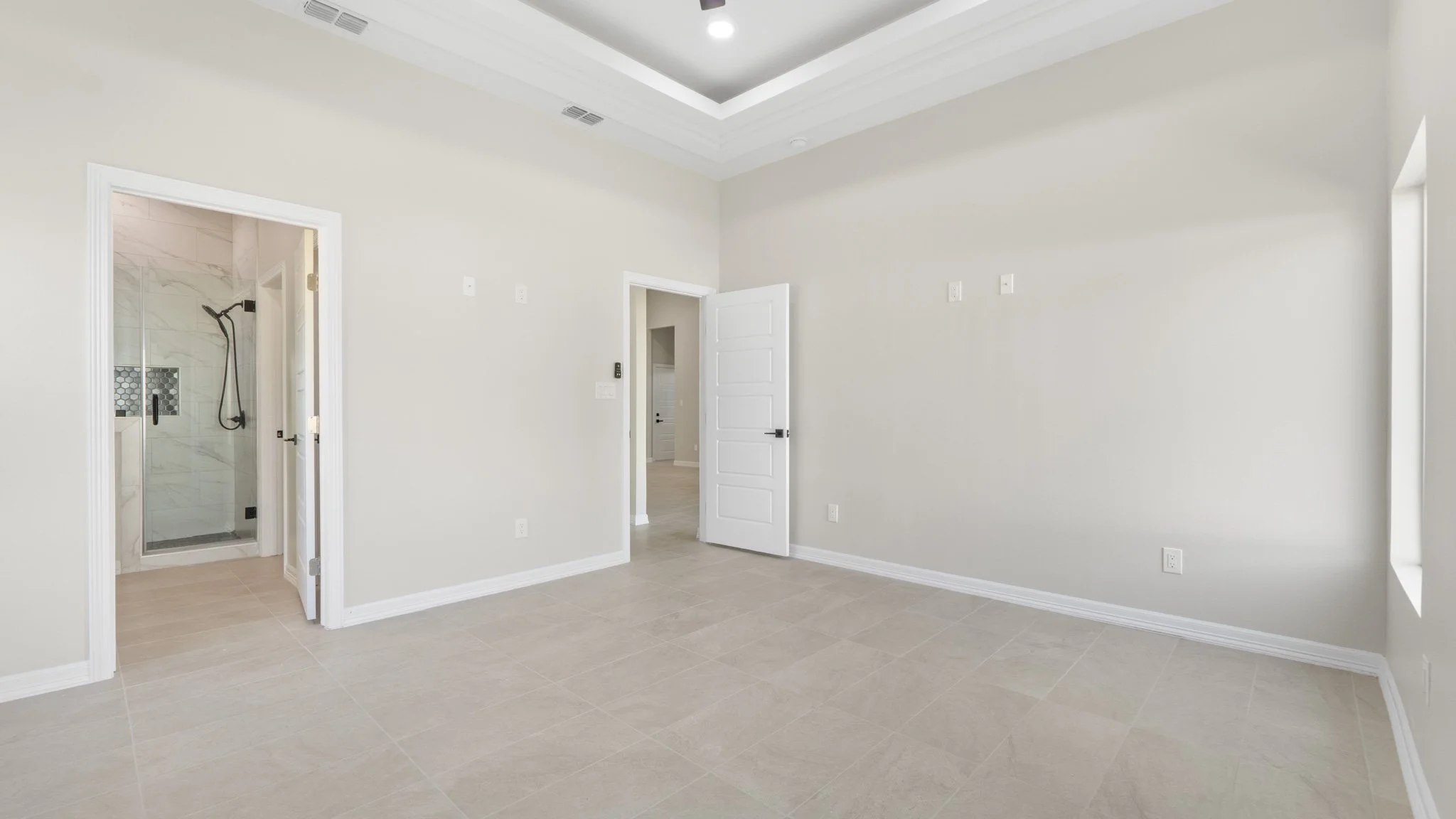 Habitación vacía con paredes blancas, suelo de baldosas beige, puerta abierta a baño con ducha y una ventana pequeña a la derecha.