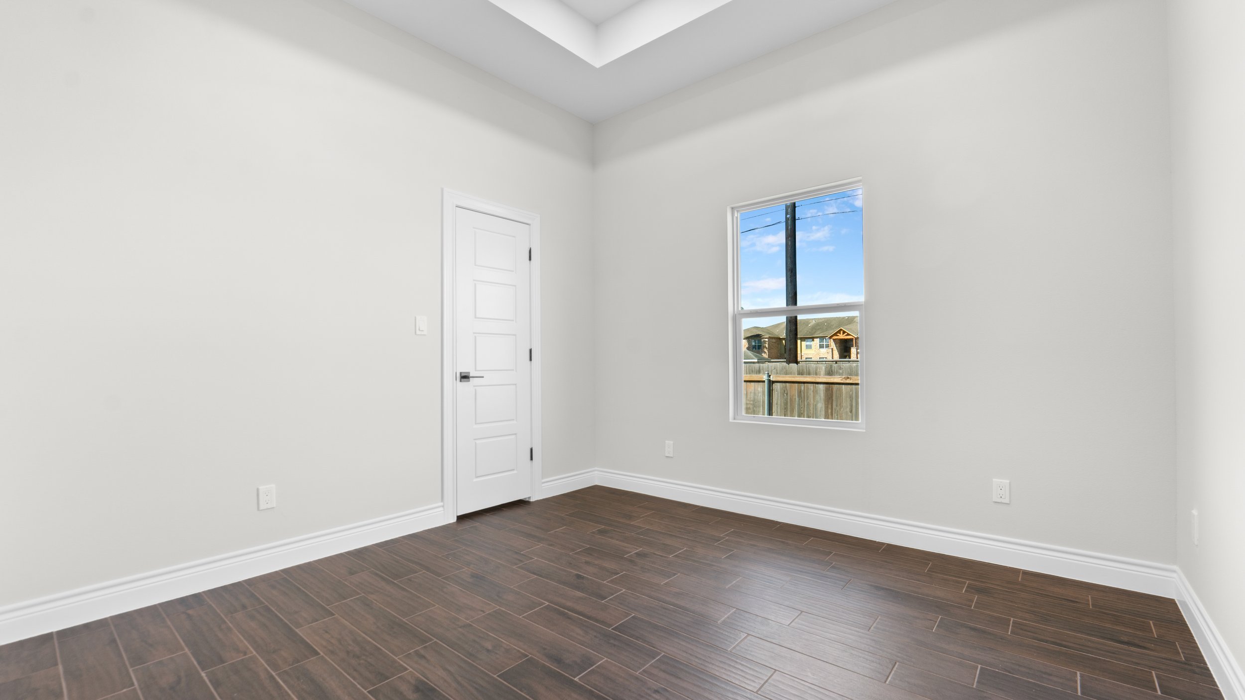 Habitación vacía con paredes blancas, ventana con vista a un vecindario y piso de madera