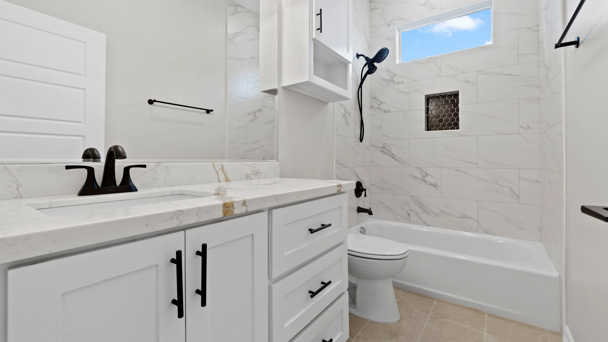 Baño moderno con ducha, lavabo con encimera de mármol y armario blanco, inodoro, ventana pequeña y detalles en negro en accesorios.