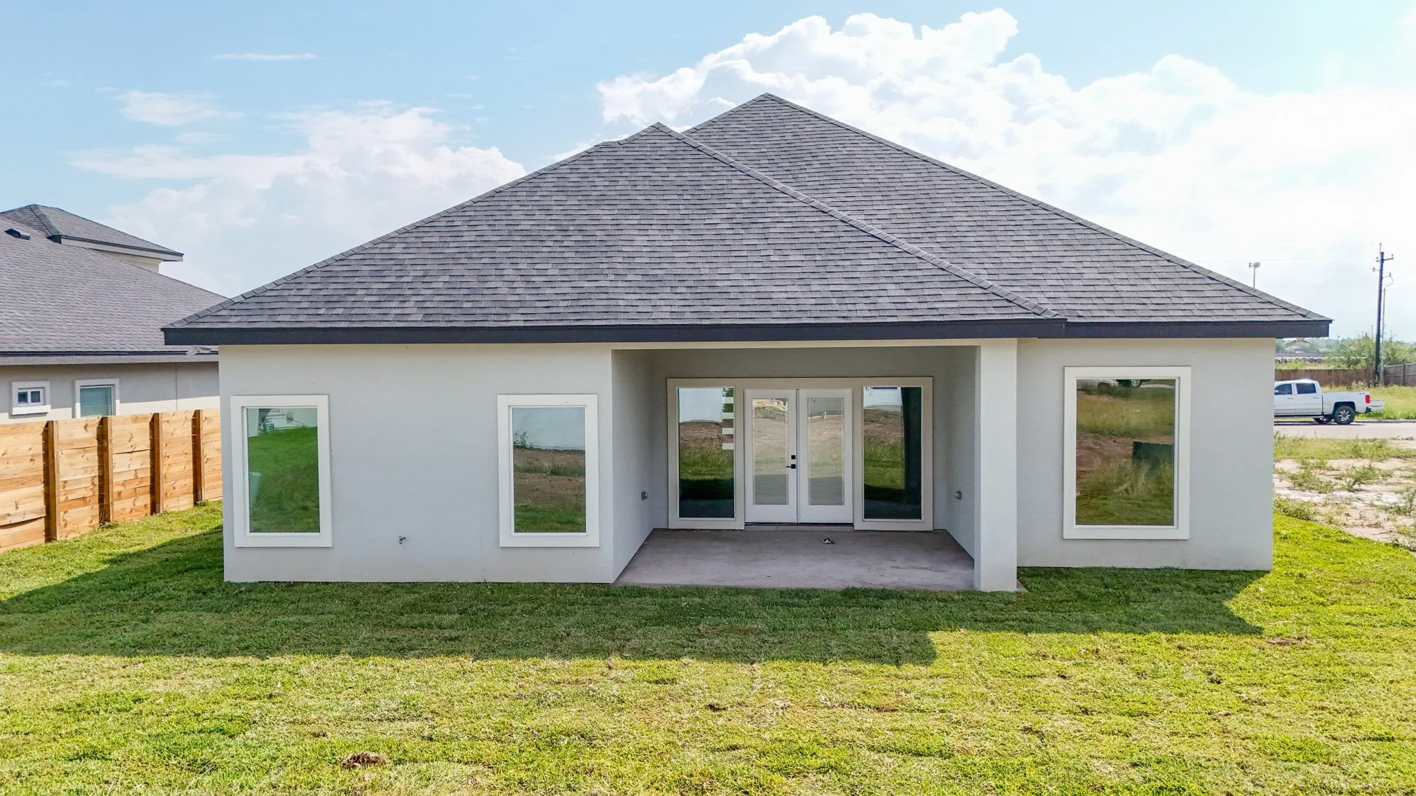 Casa moderna con paredes blancas, ventanas cuadradas, puerta de vidrio y un techo de tejas grises en un patio con césped y cielo con nubes