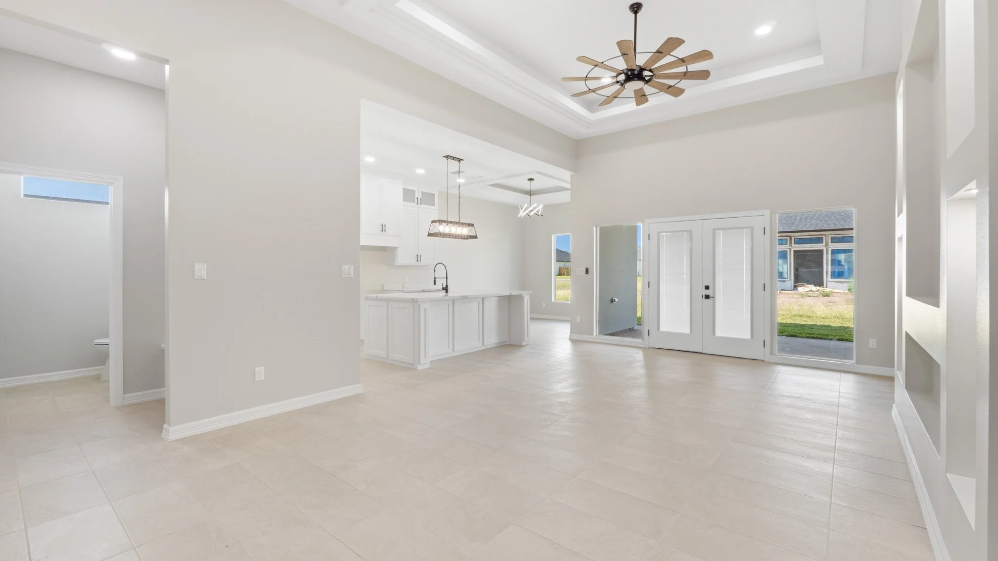Interior de una sala de estar moderna con puertas de vidrio, techo con iluminación empotrada y ventilador de techo de diseño en madera. Cocina blanca en el fondo con isla y ventanas que dejan pasar luz natural.