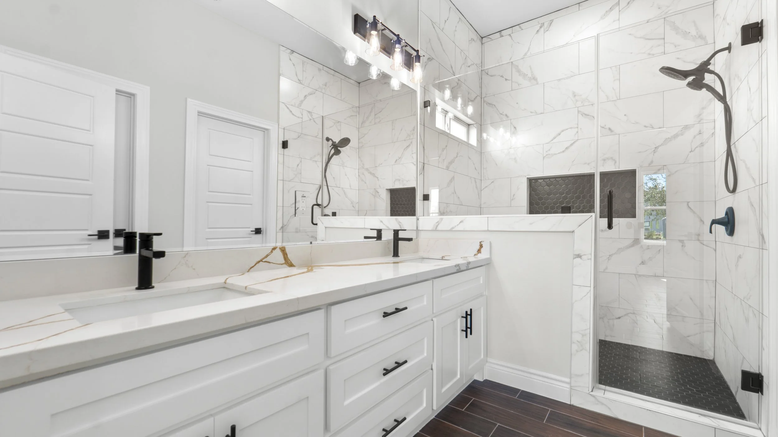 Baño con doble lavabo de mármol blanco, gabinetes blancos, grifos negros, pared de azulejos blancos con vetas grises, ducha con puertas de vidrio, azulejos blancos y pequeños nichos de almacenamiento, ventanas pequeñas, iluminación con bombillas expuestas.