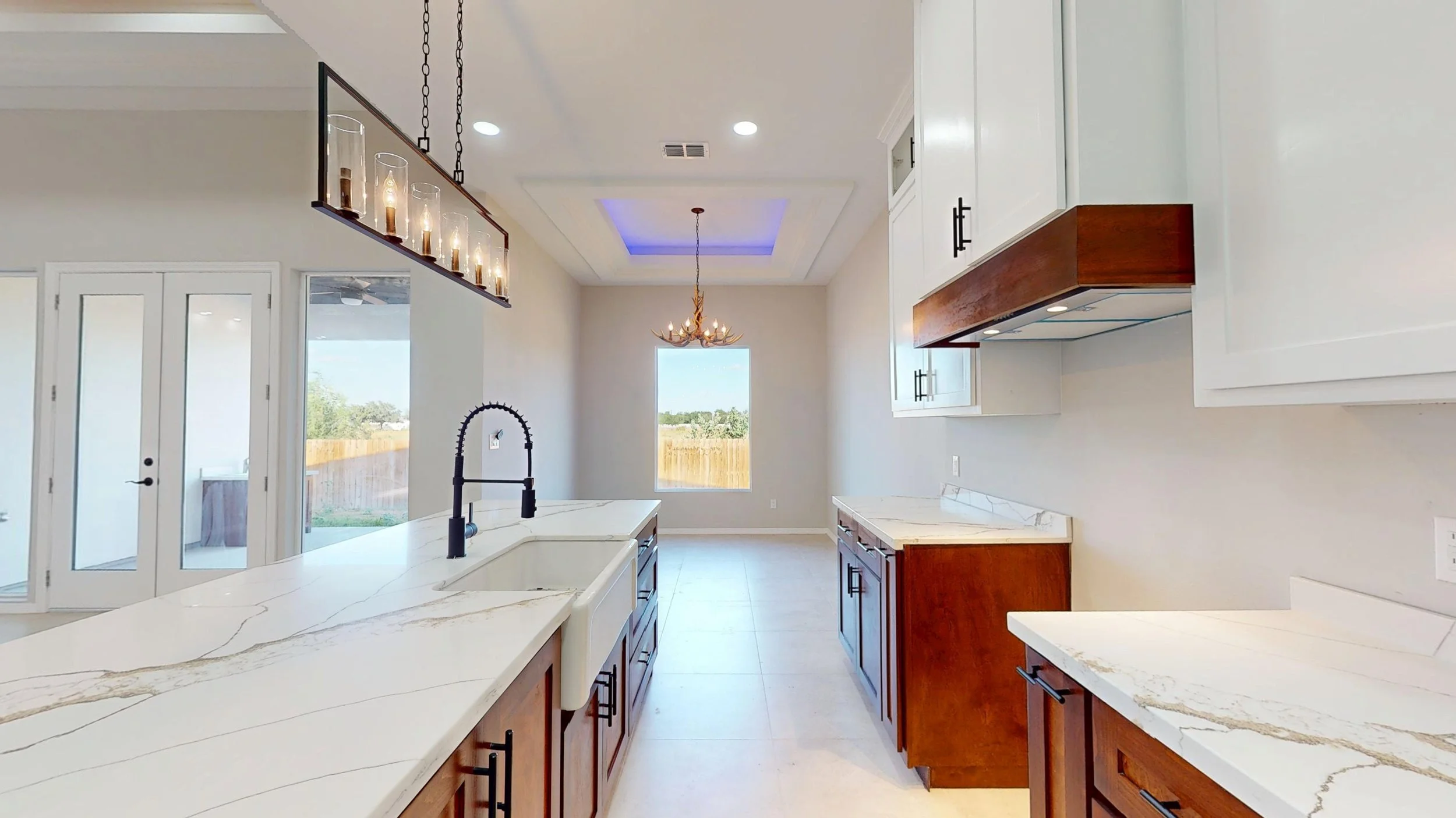 Cocina moderna con isla central, barandales de madera, gabinetes blancos, encimera de mármol, fregadero con grifo negro, ventanas grandes y puertas de vidrio, iluminación empotrada y candelabro decorativo.