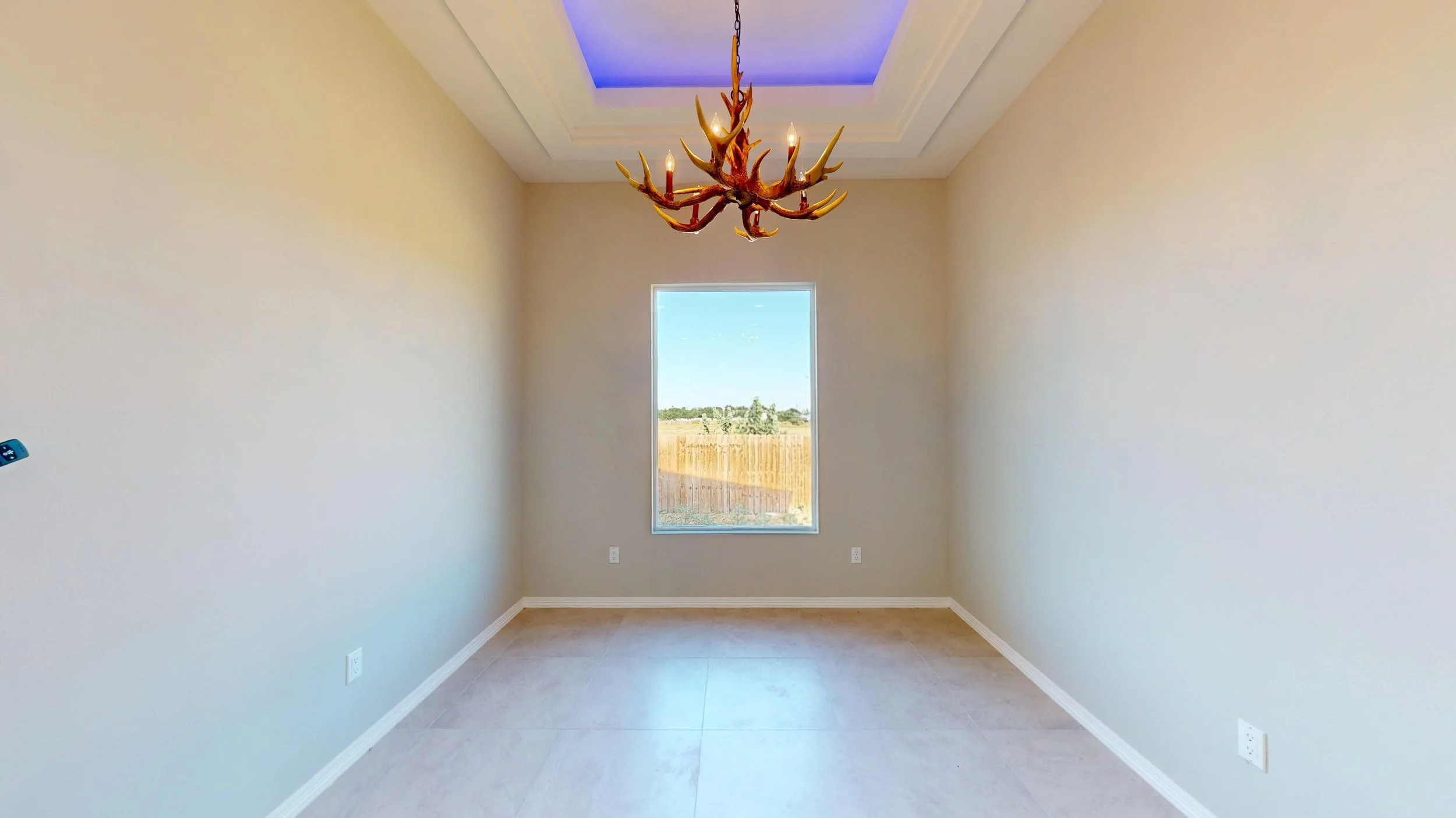Habitación vacía con paredes beige, una ventana que muestra un espacio exterior con césped y árboles, un candelabro decorativo en forma de cornamenta de ciervo en el techo, y piso de baldosas claras.