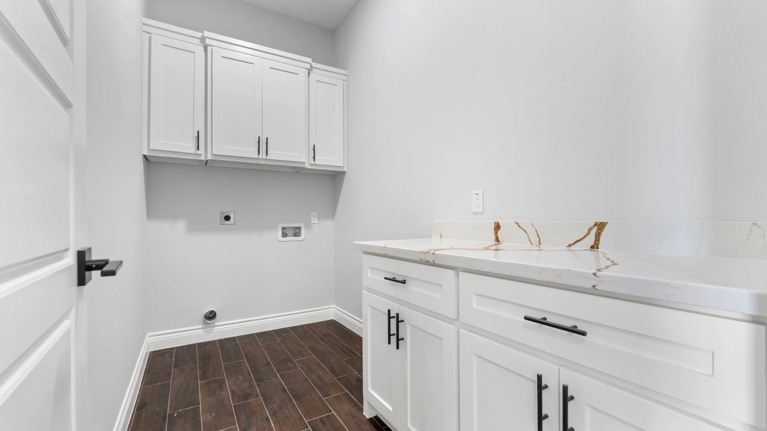 Cuarto de lavandería con gabinetes blancos, encimera de mármol y suelo de madera oscura.