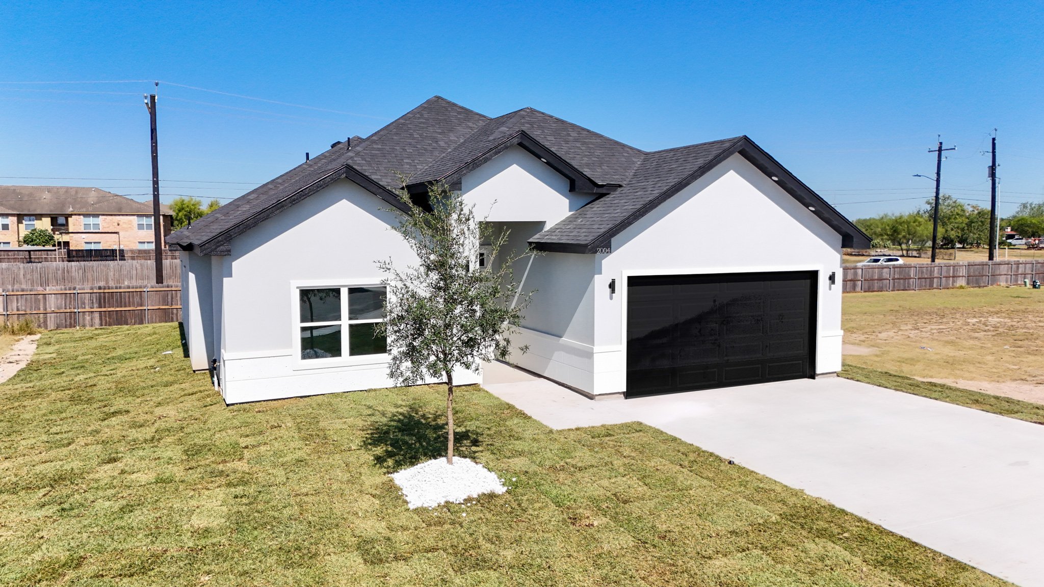 Casa moderna de paredes blancas, techo gris con varias secciones, garaje negro, pequeña cerca de árbol en jardín de césped