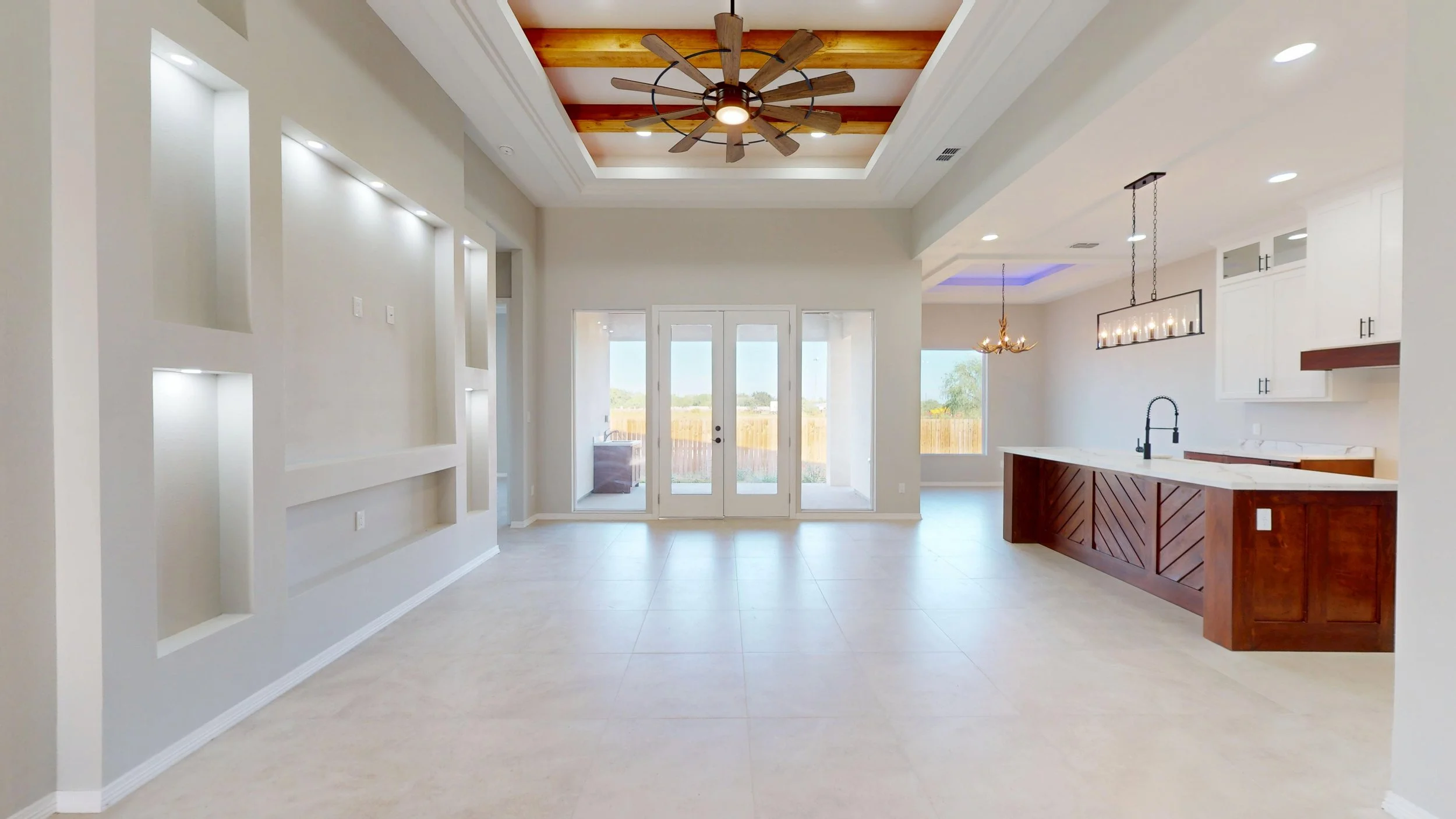 Interior de sala moderna con puertas de cristal que dan a un patio, techo de madera con ventilador de aspas, paredes blancas, espacio abierto con cocina a la derecha, con isla de madera y fregadero, lámpara colgante, y ventanas grandes que dejan entrar luz natural.