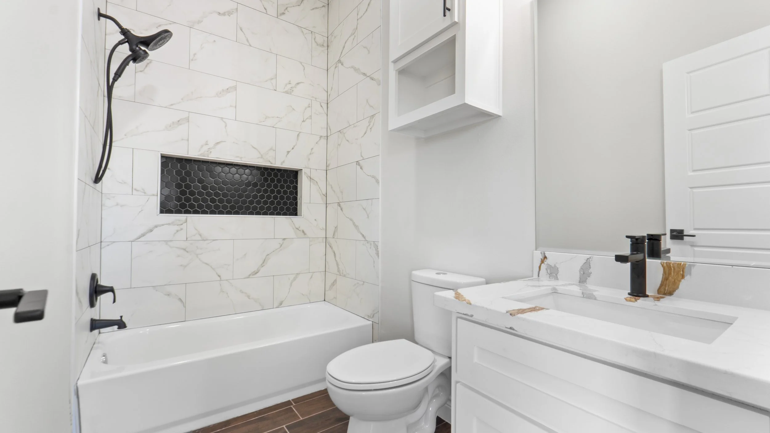 Baño moderno con ducha, inodoro y lavabo de mármol con grifo negro, azulejos blancos con vetas doradas, piso de madera y una pared con gabinetes blancos.