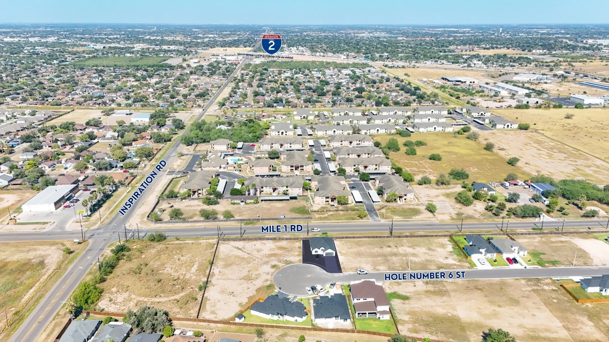 Vista aérea de un vecindario residencial con calles etiquetadas, incluyendo 'Mile 1 RD', 'Inspiration Rd' y 'Hole Number 6 St'. Se observan casas, áreas verdes y expansión urbana, con una autopista de interés llamada 'Interstate 2' en el fondo.
