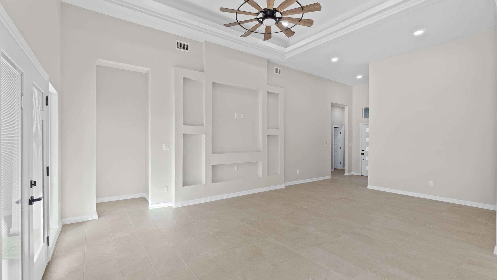 Sala de estar vacía con paredes blancas, suelo de baldosas beige, techo con falso cielo y ventilador de techo de madera. Entrada en la esquina izquierda y puerta en la parte derecha.