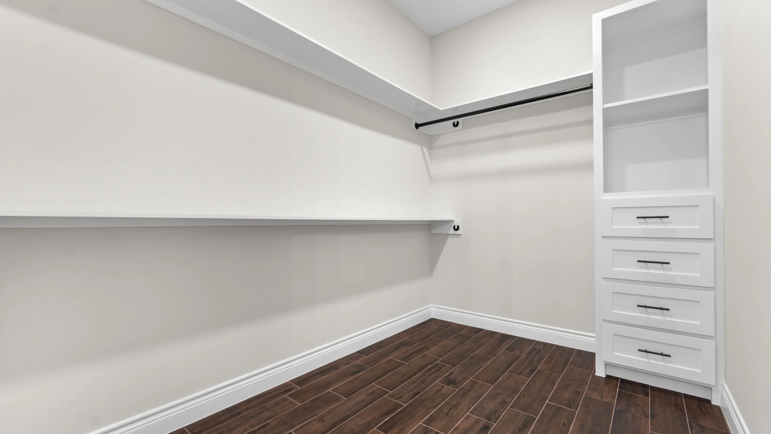 Vestidor con paredes blancas, moldura de base blanca, armario con estantes y gavetas, piso de madera oscura.