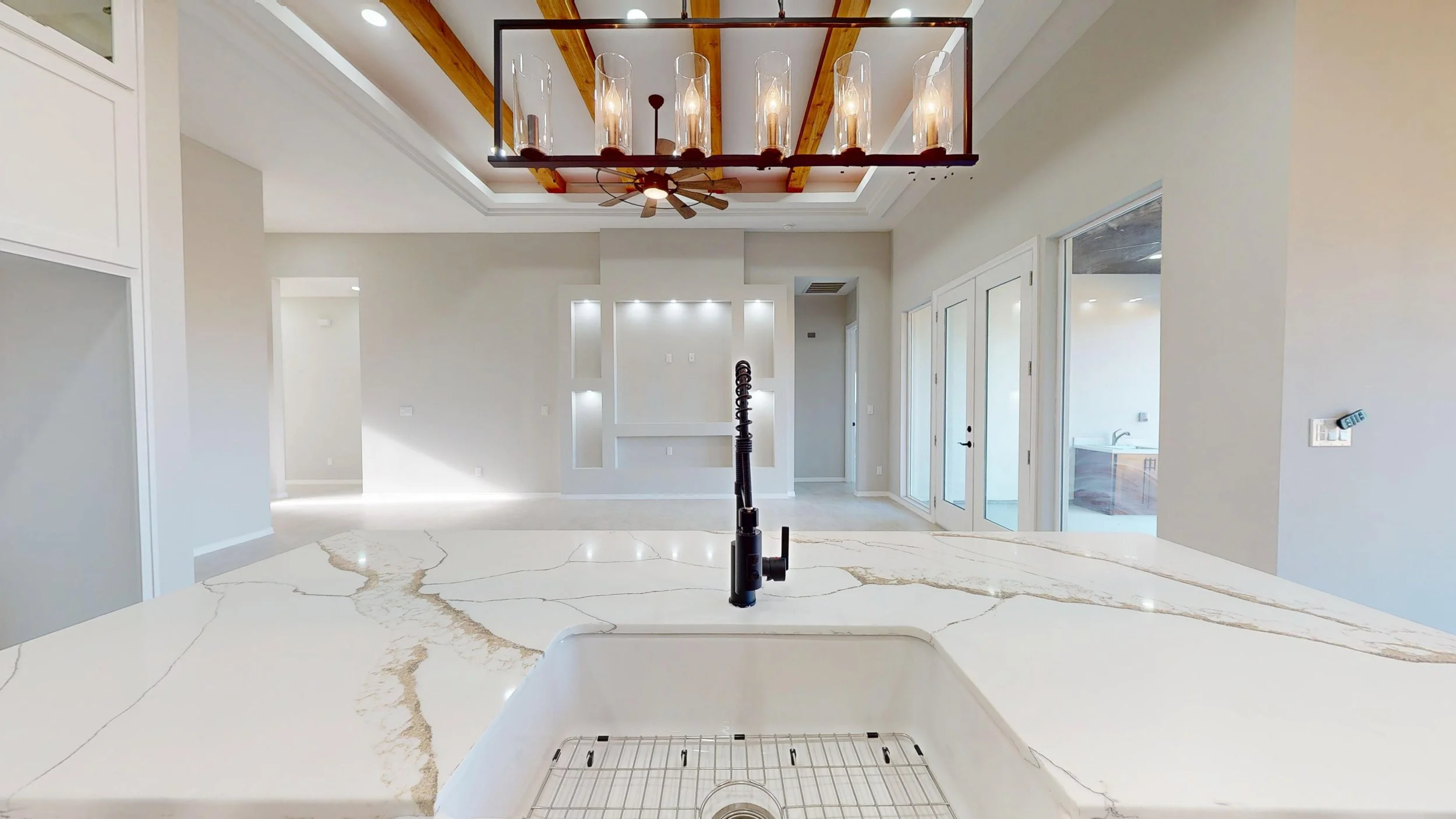 Vista desde la encimera de una cocina moderna con isla de encimera de cuarzo blanco con vetas doradas, fregadero y grifo negro, en un espacio luminoso y abierto con puertas de vidrio y paredes blancas.