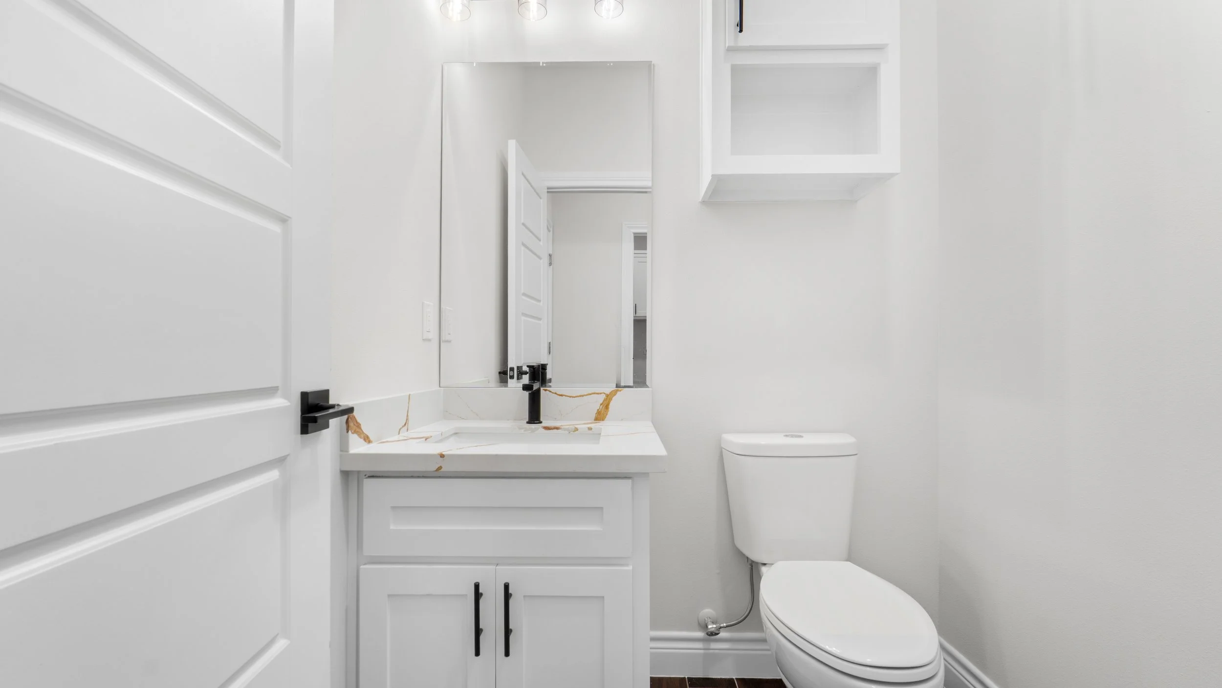 Baño con paredes blancas, lavabo con encimera de mármol con vetas doradas, espejo y sanitario, gabinete blanco, y estantes empotrados blancos.