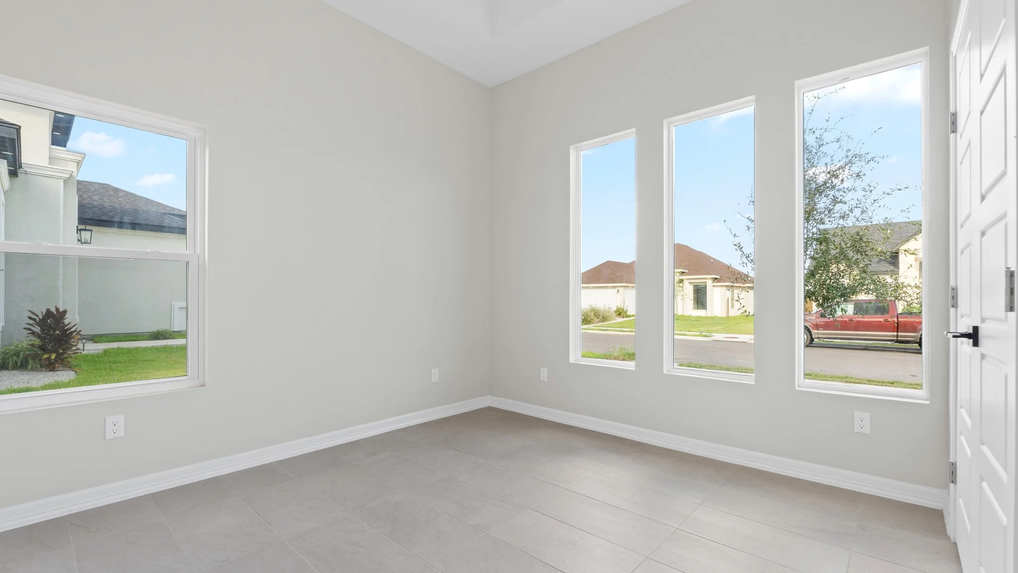 Habitación vacía con paredes blancas, ventanas grandes, suelo de baldosas y puerta blanca
