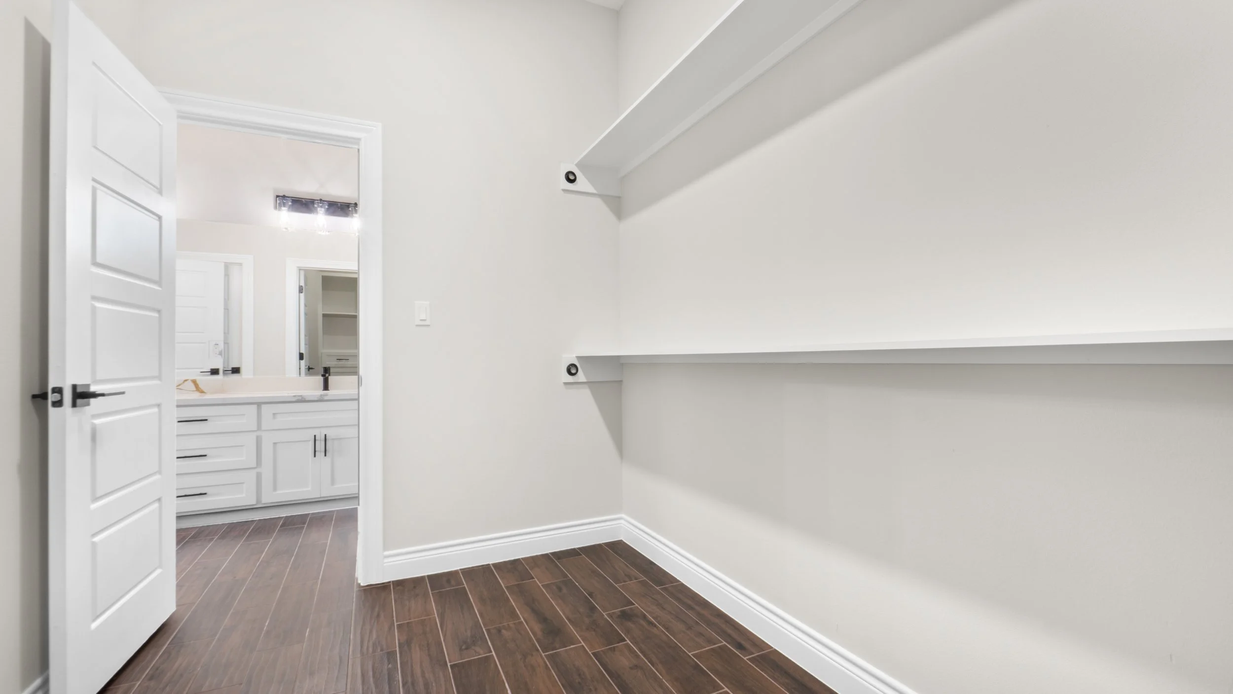 Habitación vacía con estantes blancos en la pared derecha y suelo de madera oscura, puerta abierta a un baño con lavabos blancos y espejo, en un entorno moderno y limpio.