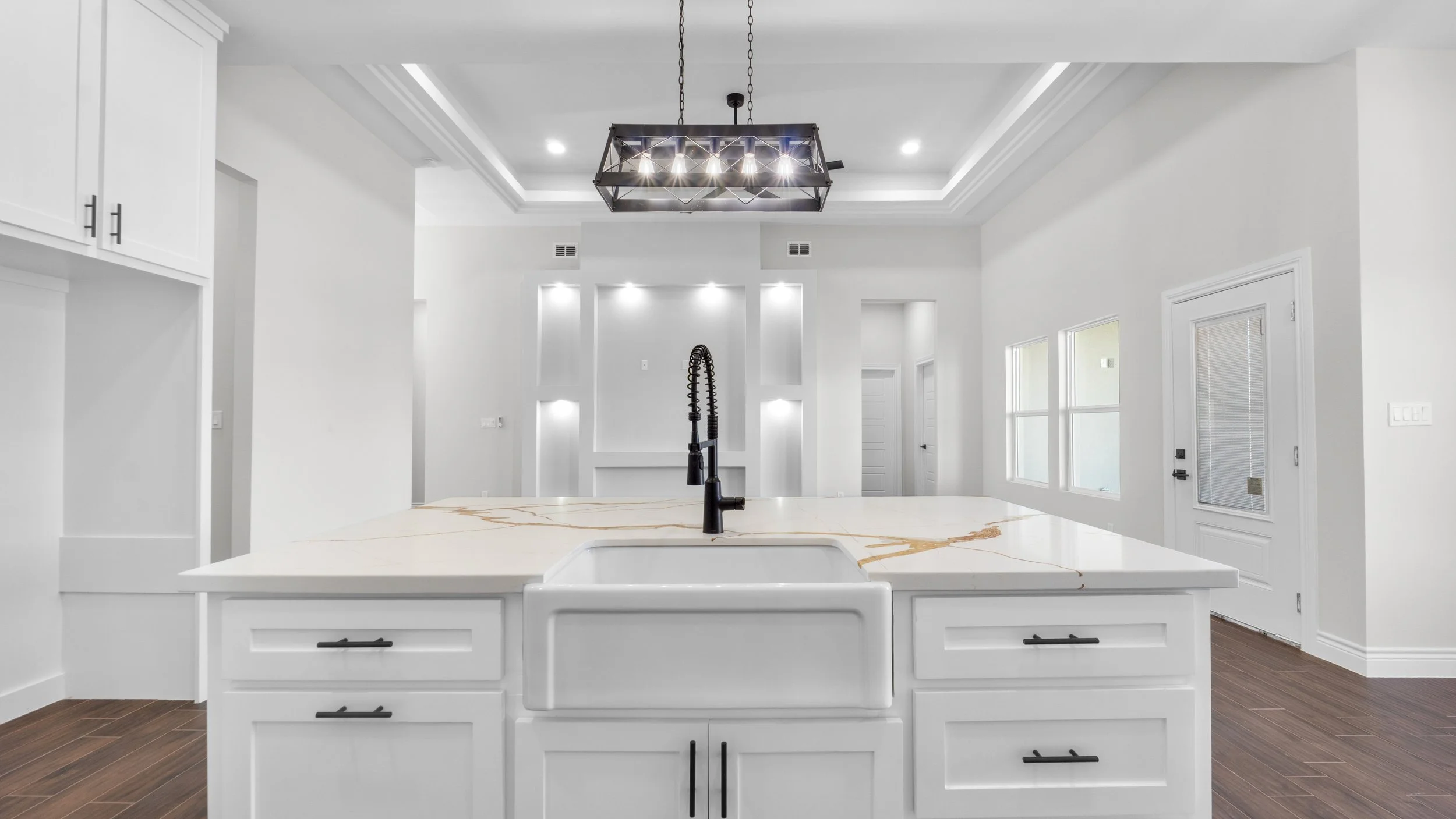 Cocina moderna con encimera de mármol blanca, fregadero, grifo negro, iluminación empotrada y ventana grande en pared blanca, en un espacio abierto con piso de madera oscura.