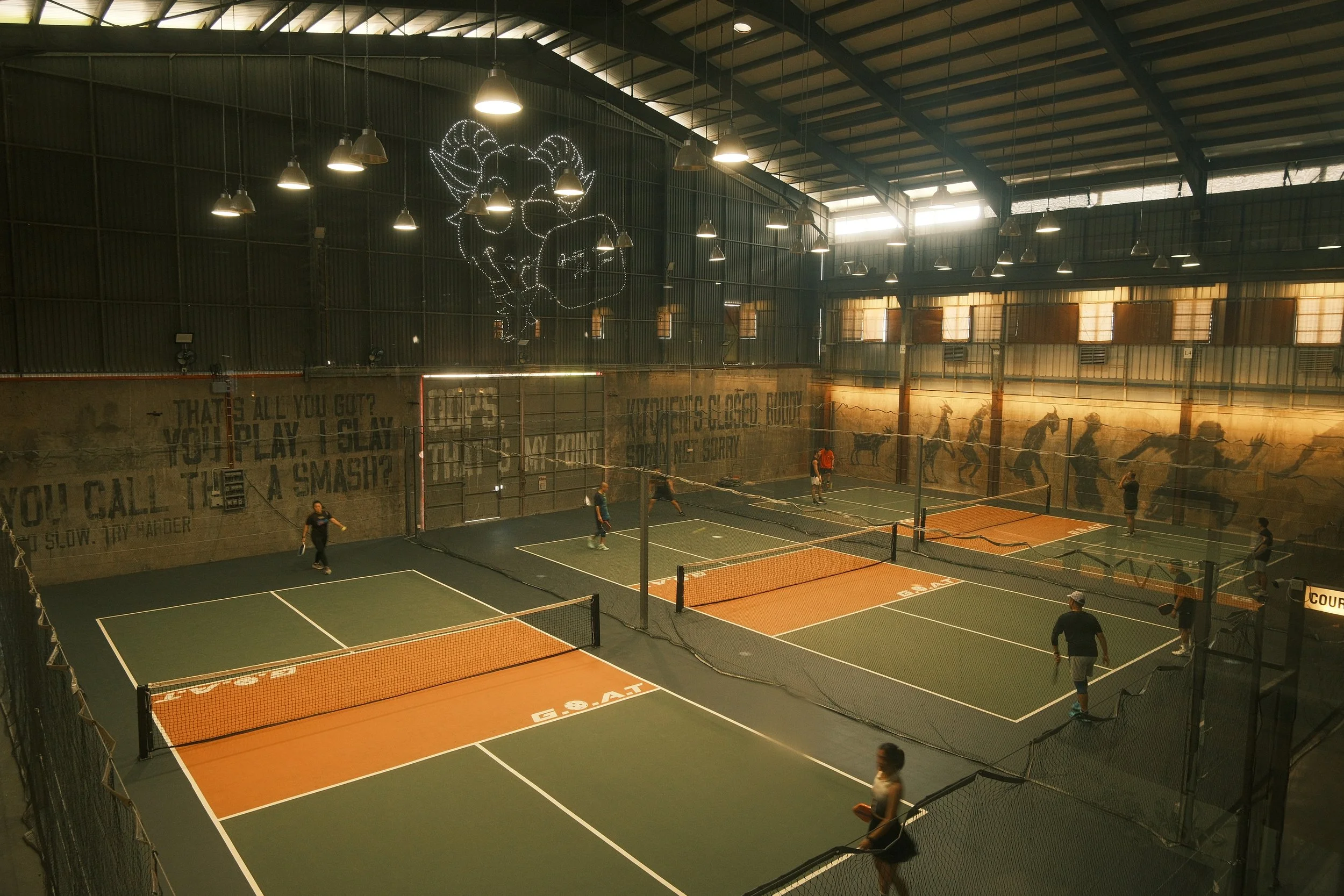 Indoor tennis facility with multiple tennis courts, players practicing, graffiti-style murals on the walls, and a large decorative light fixture on the ceiling.
