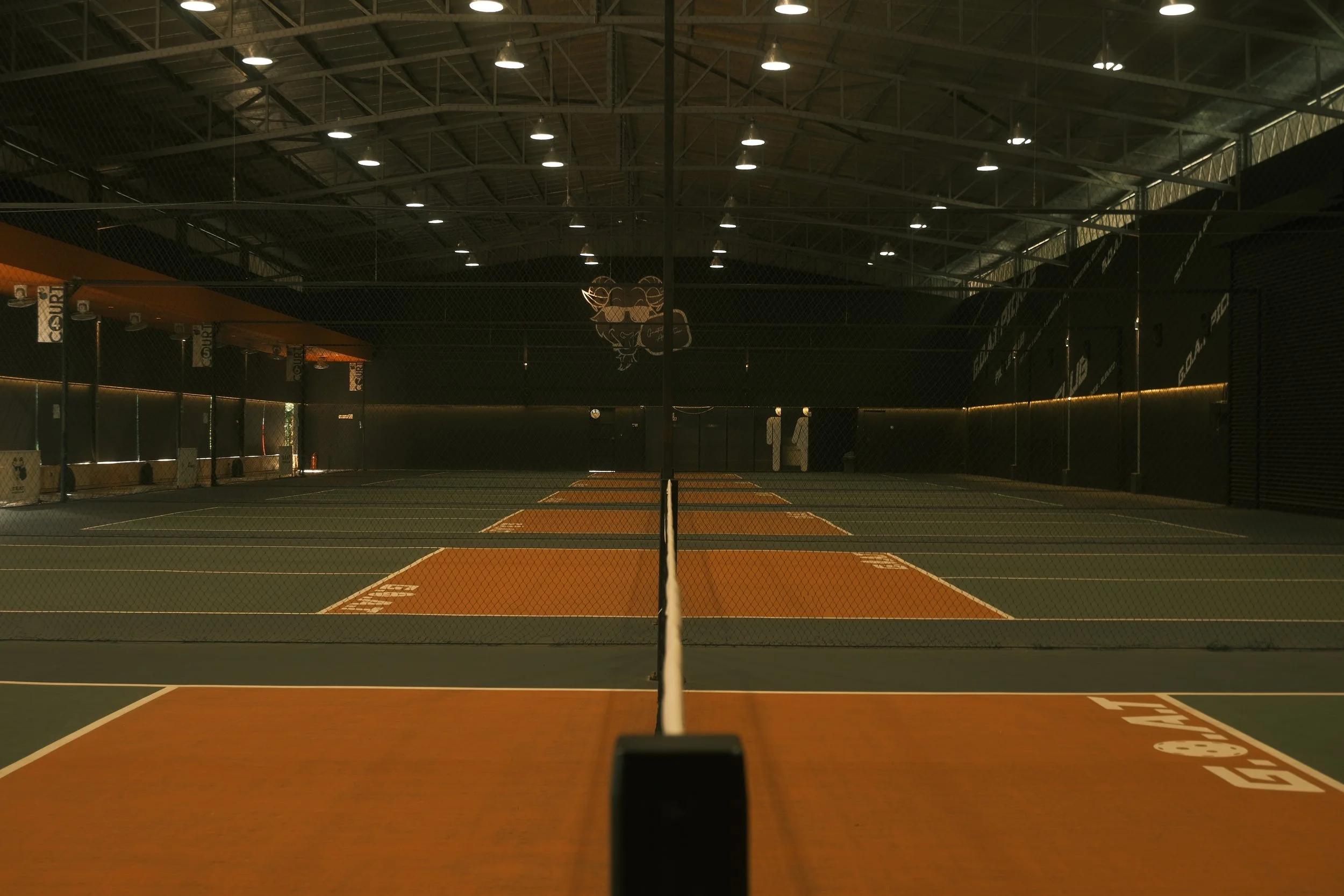 Empty indoor pickleball court with orange and green surfaces, illuminated by overhead lights, with a net in the center and black walls surrounding the court.