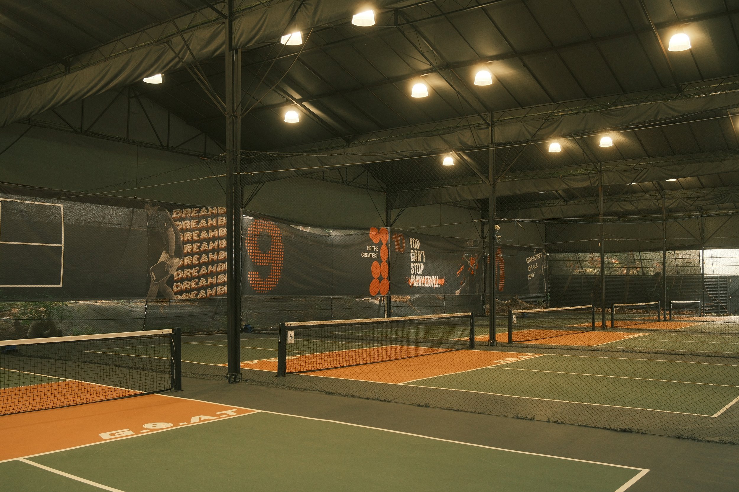 Indoor tennis courts with multiple courts separated by black nets, under a high ceiling with overhead lighting.