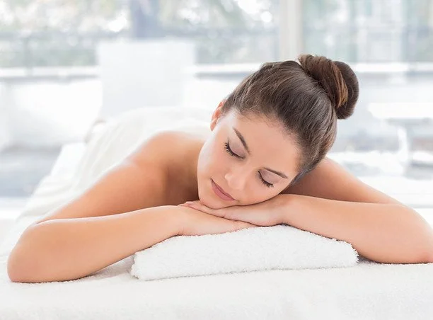 A woman with her hair tied back resting her head on a folded towel, lying face down on a massage table in a well-lit room.