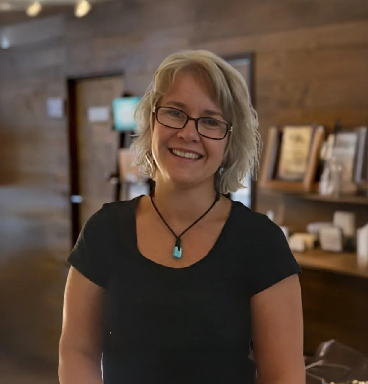A smiling woman with glasses and shoulder-length blonde hair, wearing a black shirt and a turquoise pendant necklace, standing indoors with wooden walls and shelves in the background.