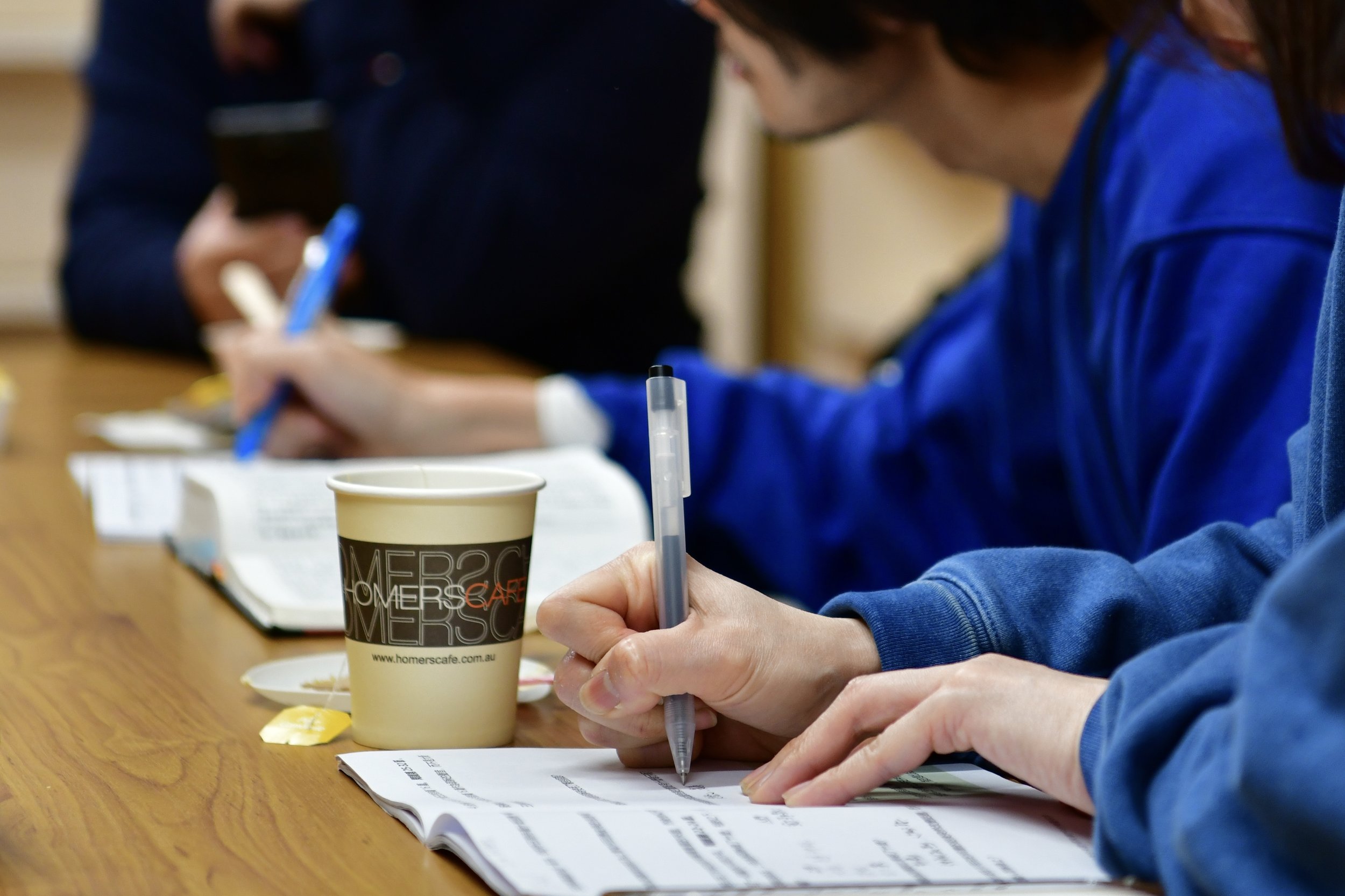 A close-up shot of the hands of two people as they sit at a table, writing in a booklet.