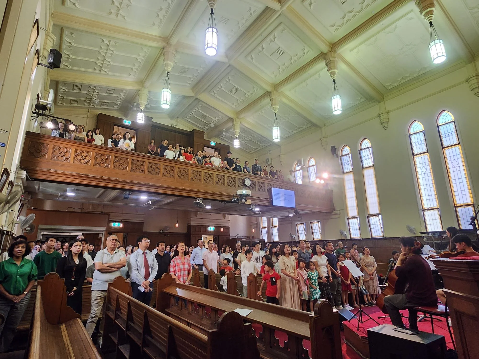 A large group of people inside a church, standing and singing, with a musician playing guitar at the front. The church has high arched windows, chandeliers, and wooden interior accents. Some people are seated in the pews, while others are on a balcony.