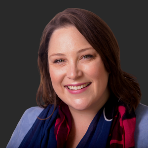 Image of Johanna Kohler from Compelling Copy. She is a woman with shoulder-length brown hair smiling, wearing a dark scarf and a blue blazer against a dark background.