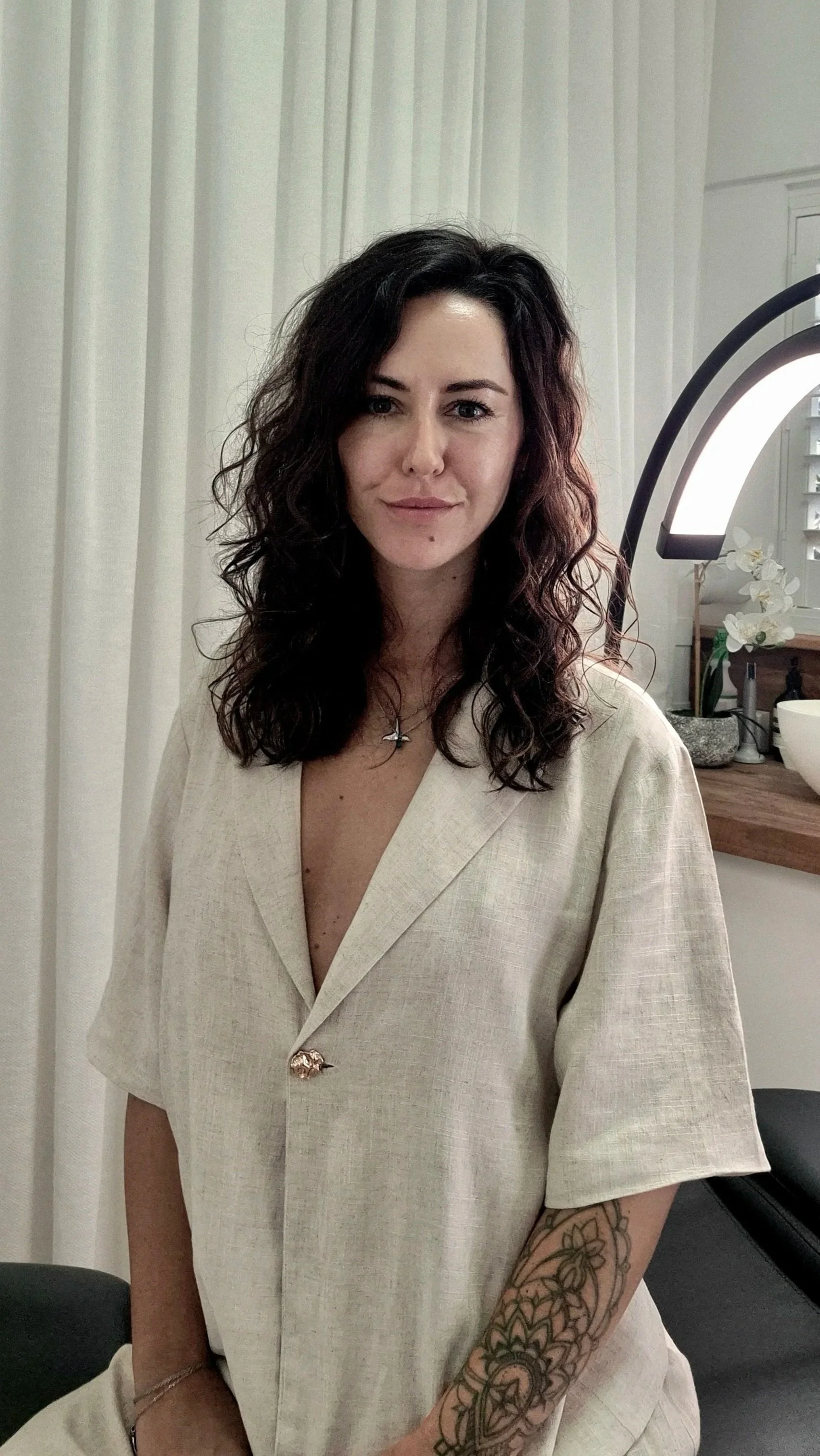 A woman with dark curly hair wearing a beige outfit and a cross necklace, standing in a room with white curtains, a wooden table with a white flower arrangement, and a large ring-shaped light.