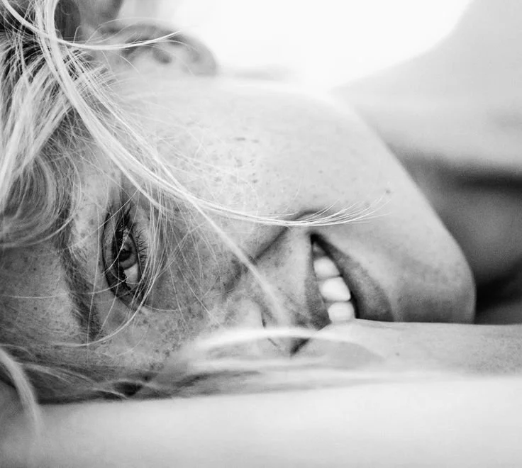 Close-up black and white photograph of a smiling woman lying down, with her face turned to the side and her hair partially covering her face.