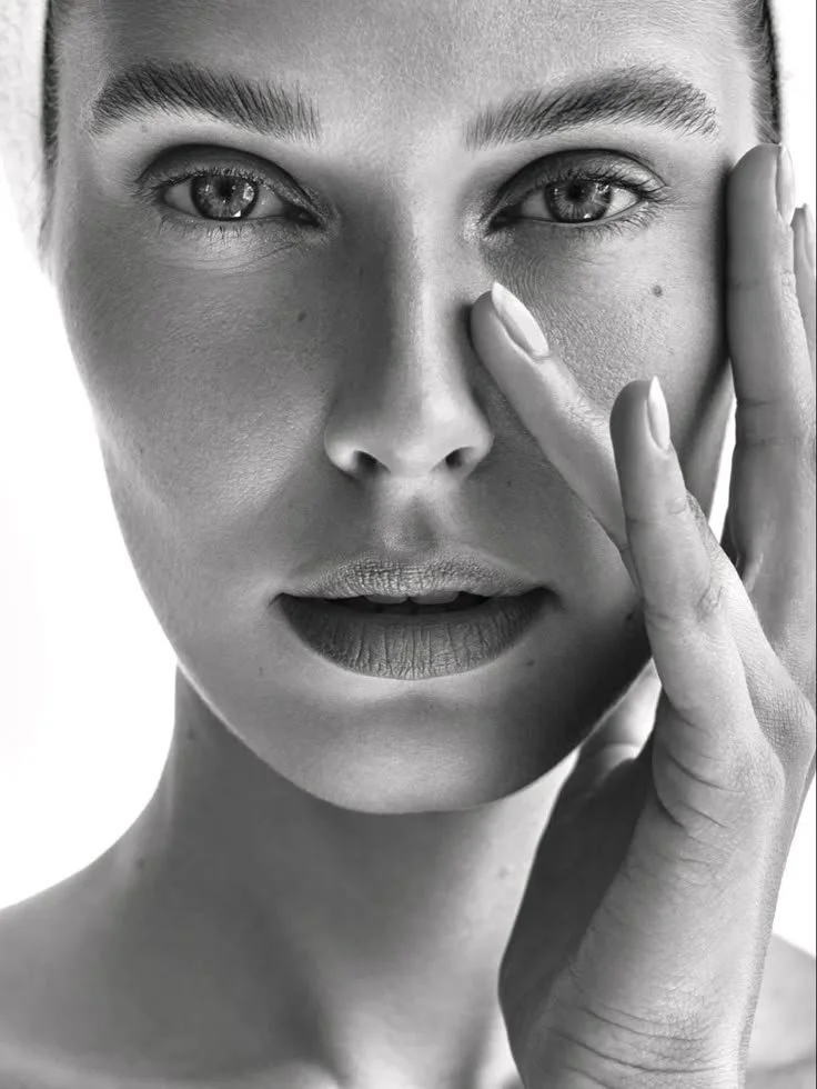 A close-up black and white photo of a woman with her hand touching her face near her nose, looking directly at the camera.