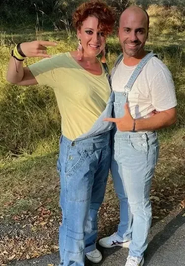 Un homme et une femme posent ensemble dans un champ, souriant, avec la femme faisant un geste de bras. La femme a les cheveux bouclés, porte un T-shirt jaune et une salopette en jean. L'homme est barbu, porte un T-shirt blanc et une salopette en jean claire.