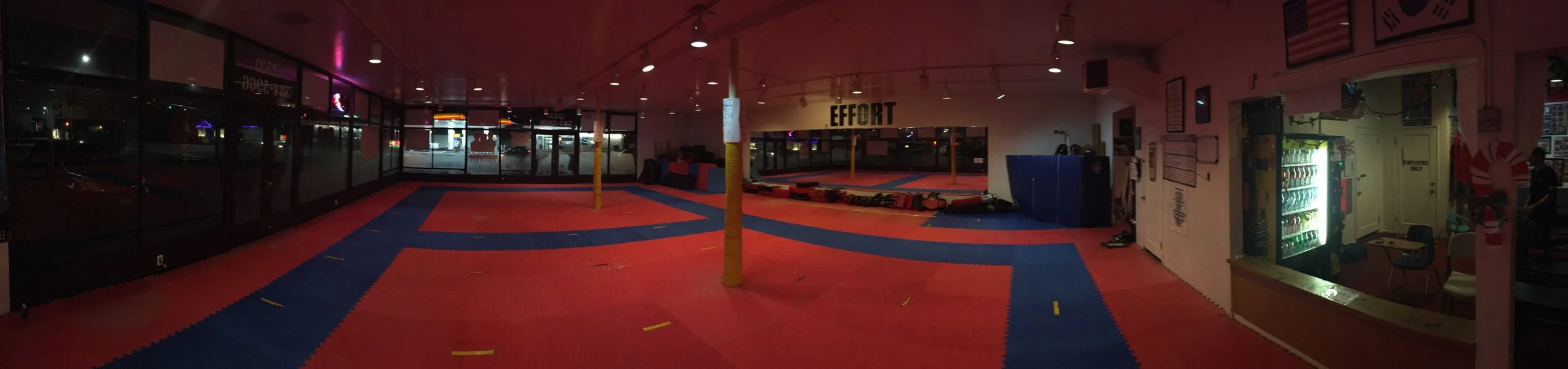 Empty martial arts training room with padded red and blue mats, yellow poles, and various training equipment. Glass windows along the left side, vending machine on the right, and a sign that reads 'EFFORT' on the back wall.