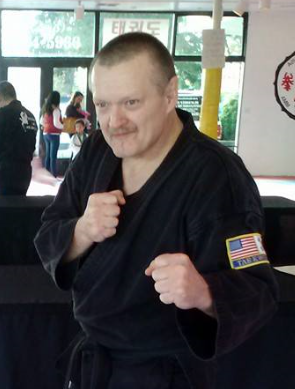 Man in black martial arts uniform with American flag patch, posing with fists raised in a martial arts stance
