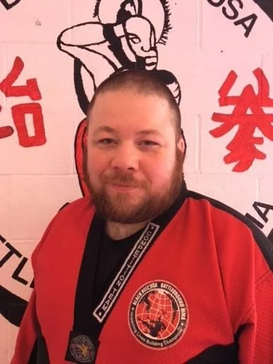 Man with a beard and short hair wearing a red martial arts gi and a black medal around his neck, standing in front of a wall with martial arts symbols and text.