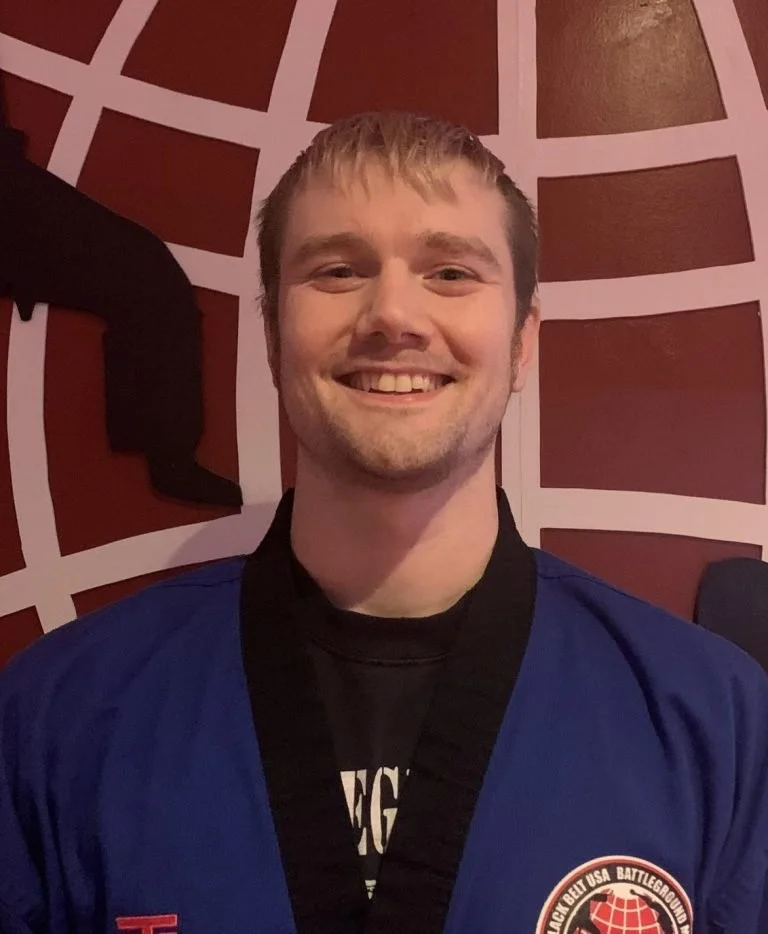 A young man smiling, wearing a blue martial arts uniform with a black belt, standing in front of a red and white geometric patterned wall.