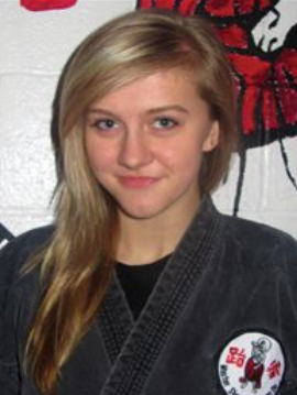 Young woman in a martial arts uniform with a patch on her chest, standing in front of a white wall with a red and black martial arts poster.