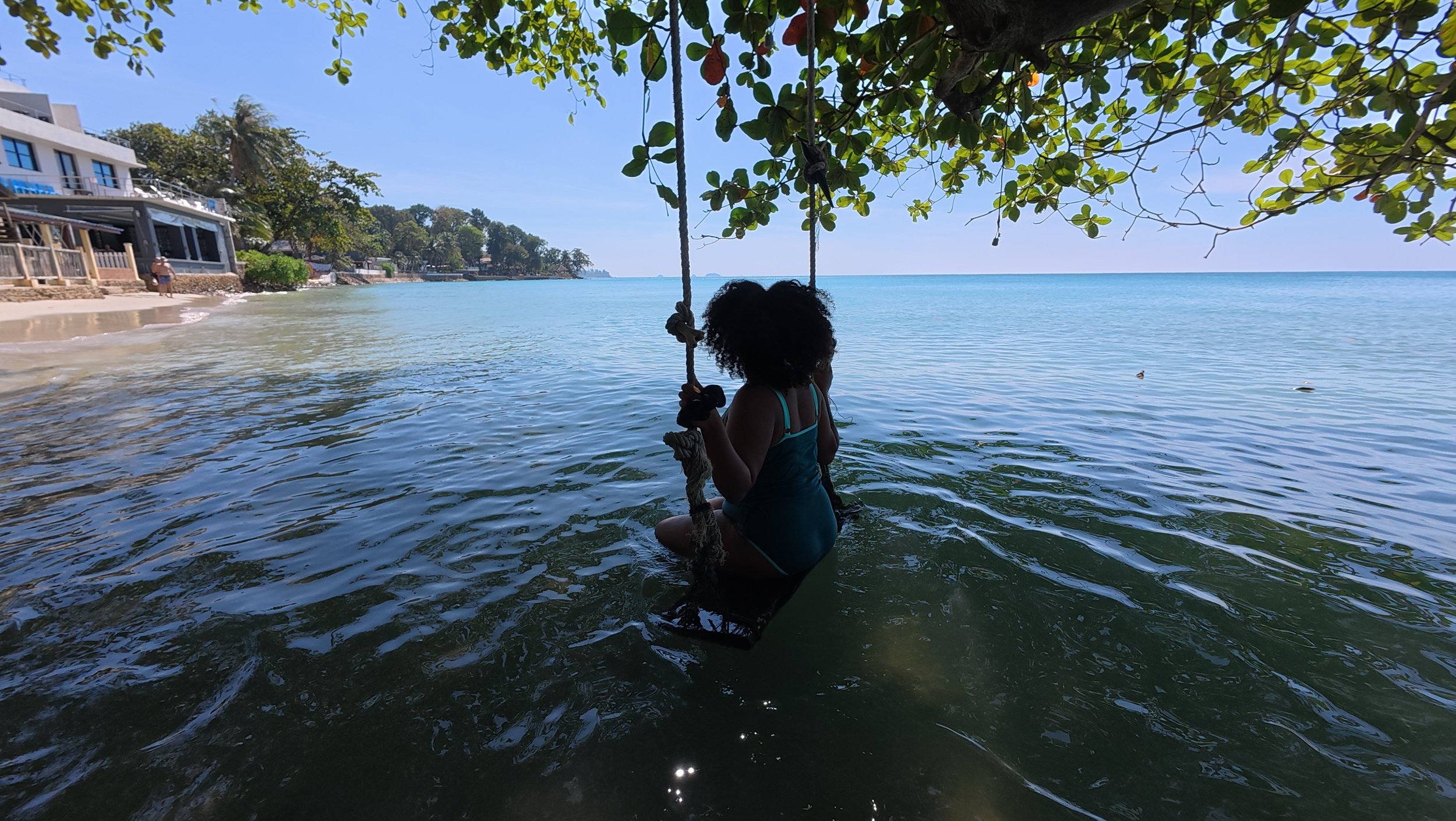 A person with curly hair sitting on a swing hanging from a tree over the water at the beach, with houses and trees along the shoreline, under a clear blue sky.