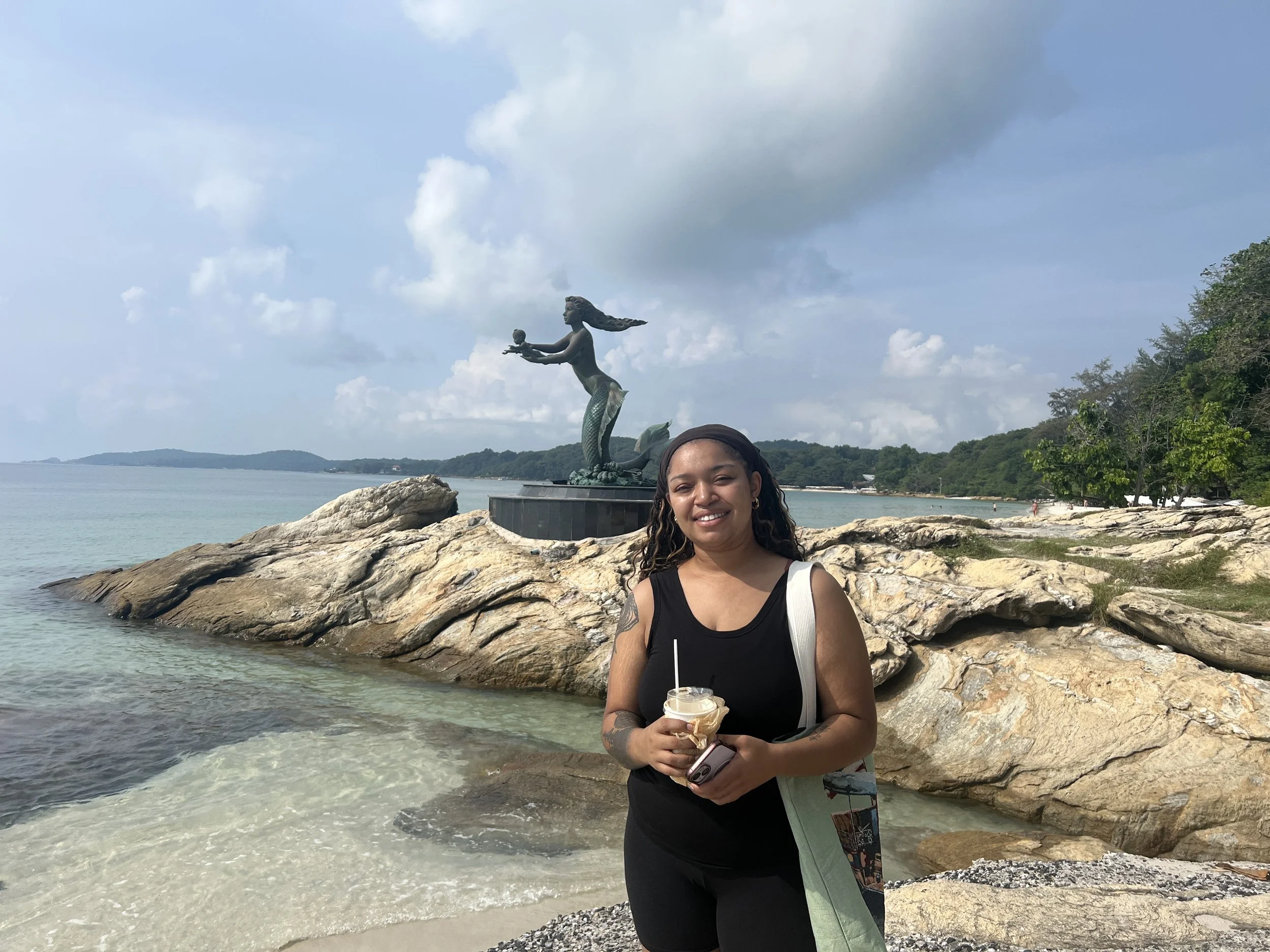 A woman holding a drink and a phone standing on rocks by the beach with a mermaid statue in the background, ocean, cloudy sky, trees on the right side.